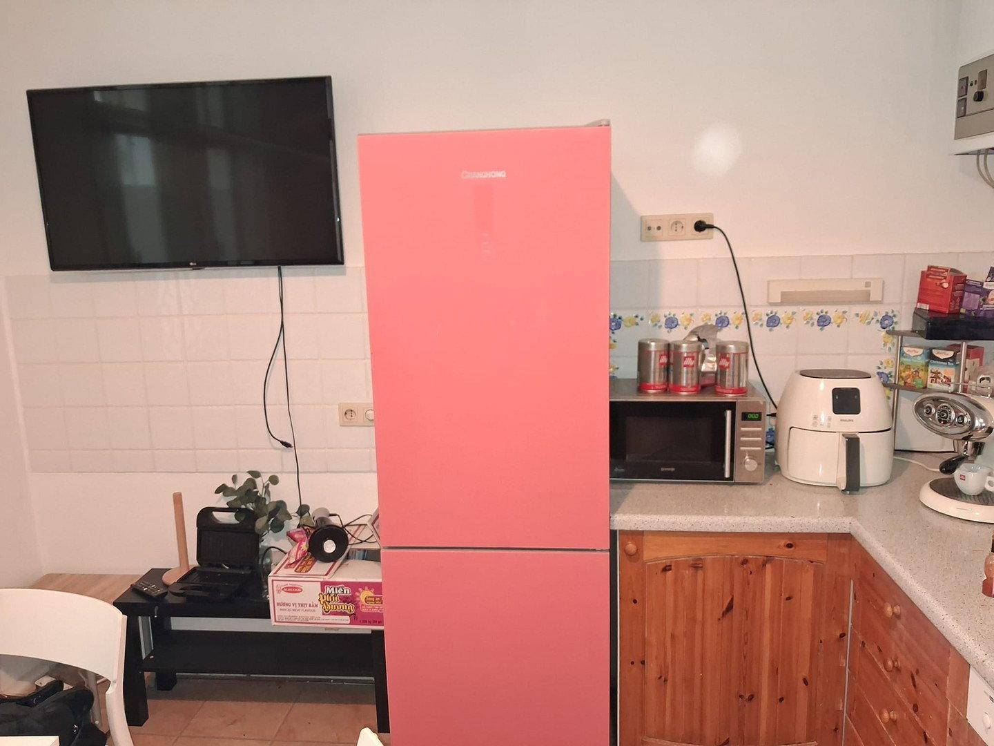 Küche mit einem auffälligen pinken Kühlschrank, Mikrowelle und einem Fernseher an der Wand.