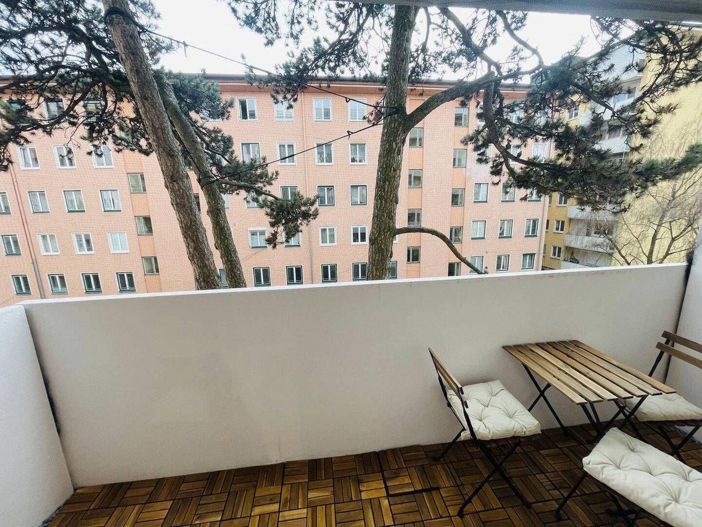 Kleiner Balkon mit Holzboden und einfacher Bestuhlung, bietet Aussicht auf die Nachbarschaft.