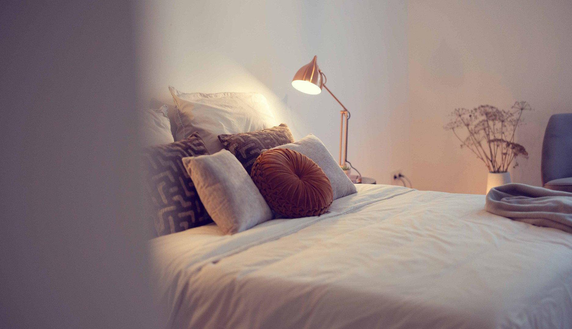 Gemütliches Bett mit vielen Kissen und warmem Licht einer modernen Nachttischlampe.