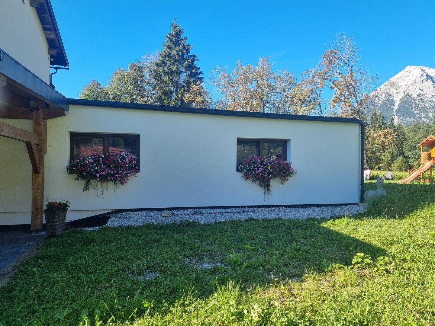 Gartenbereich mit Rasen, Blumenkästen an der Fassade und einem Spielplatz im Hintergrund.