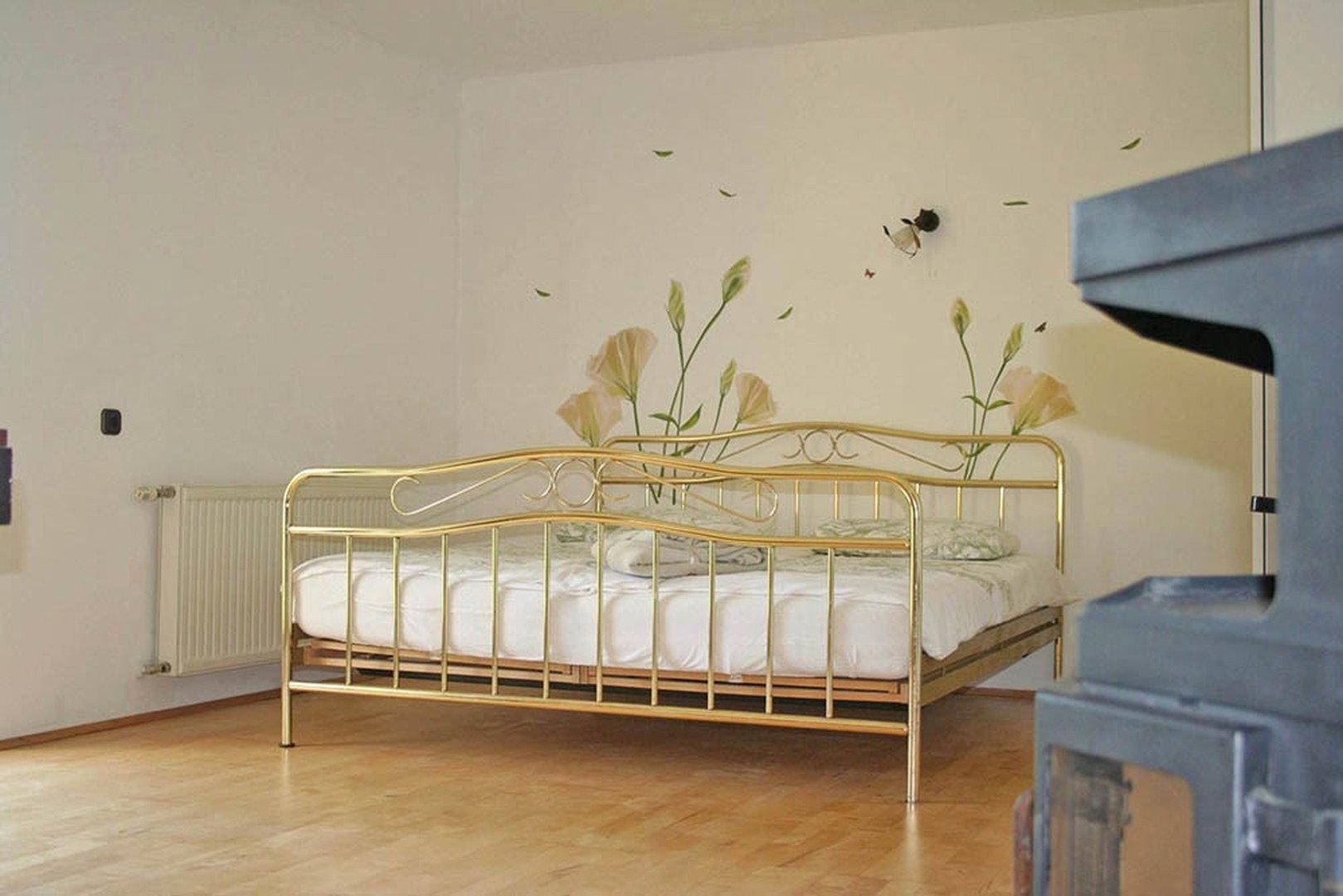Schlafzimmer mit goldenem Metallbett, Holzboden und floraler Wanddekoration.