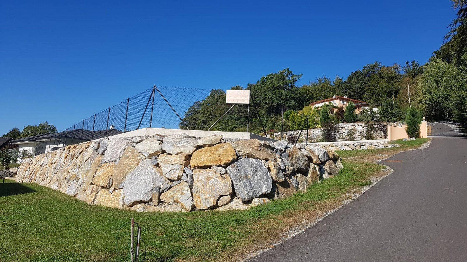 Rustikale Natursteinmauer entlang einer Zufahrt, umgeben von Grünflächen und Bäumen unter blauem Himmel.