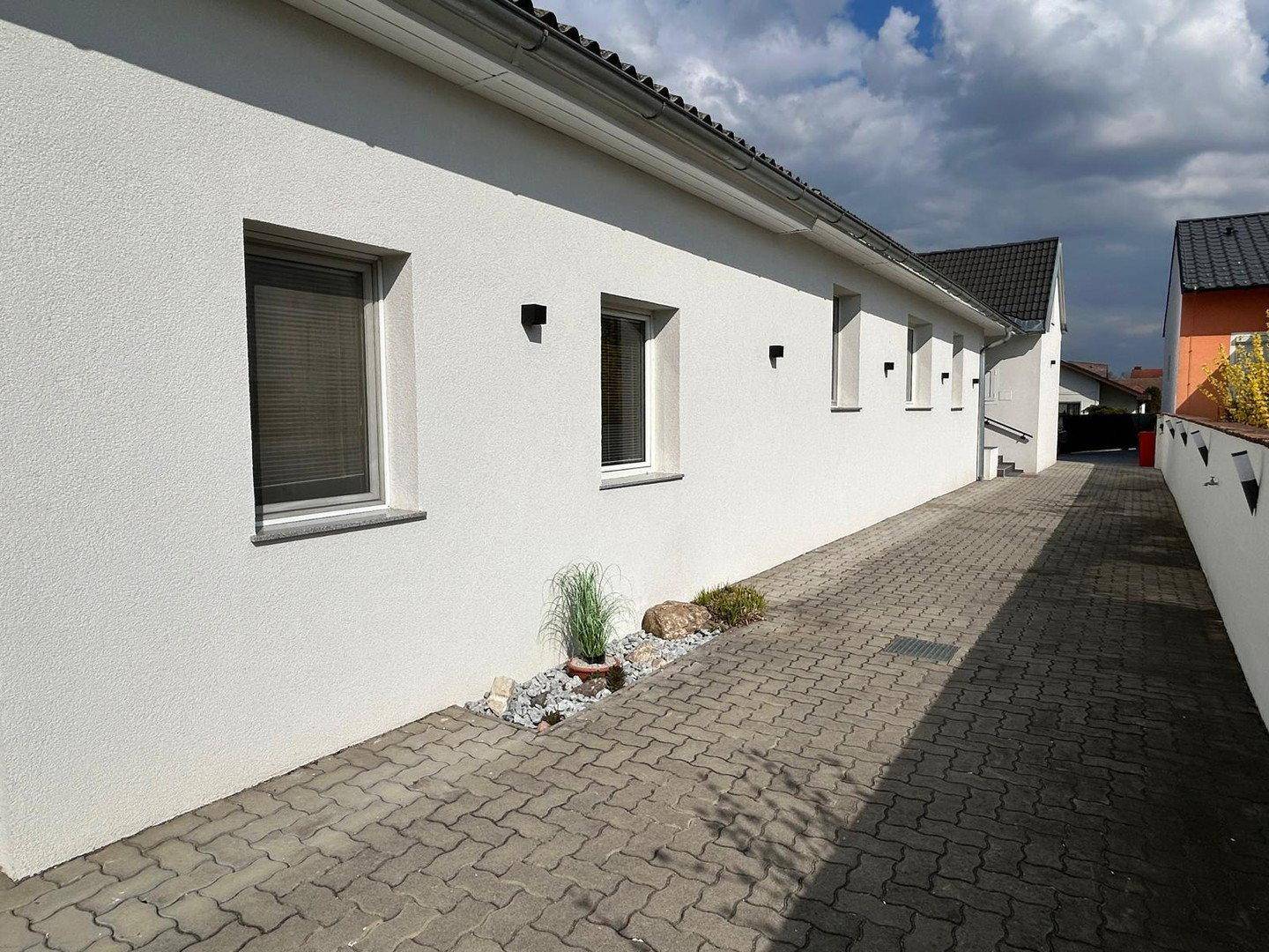 Lange, gepflasterte Zufahrt entlang der weißen Hausfassade mit kleinen Pflanzenbeeten.