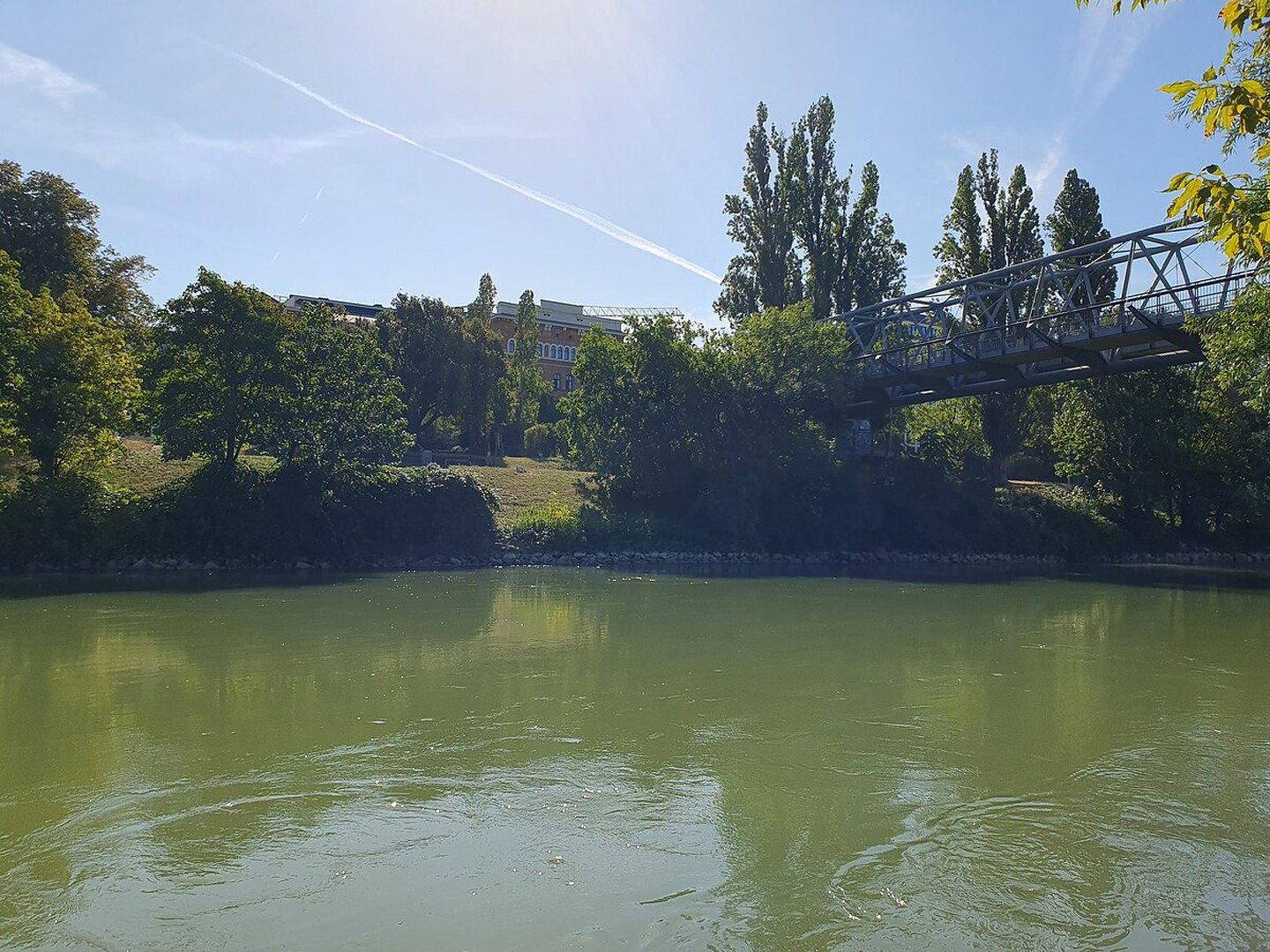 Fluss oder Kanal mit grünem Wasser, umgeben von Bäumen und einer Metallbrücke im Hintergrund.