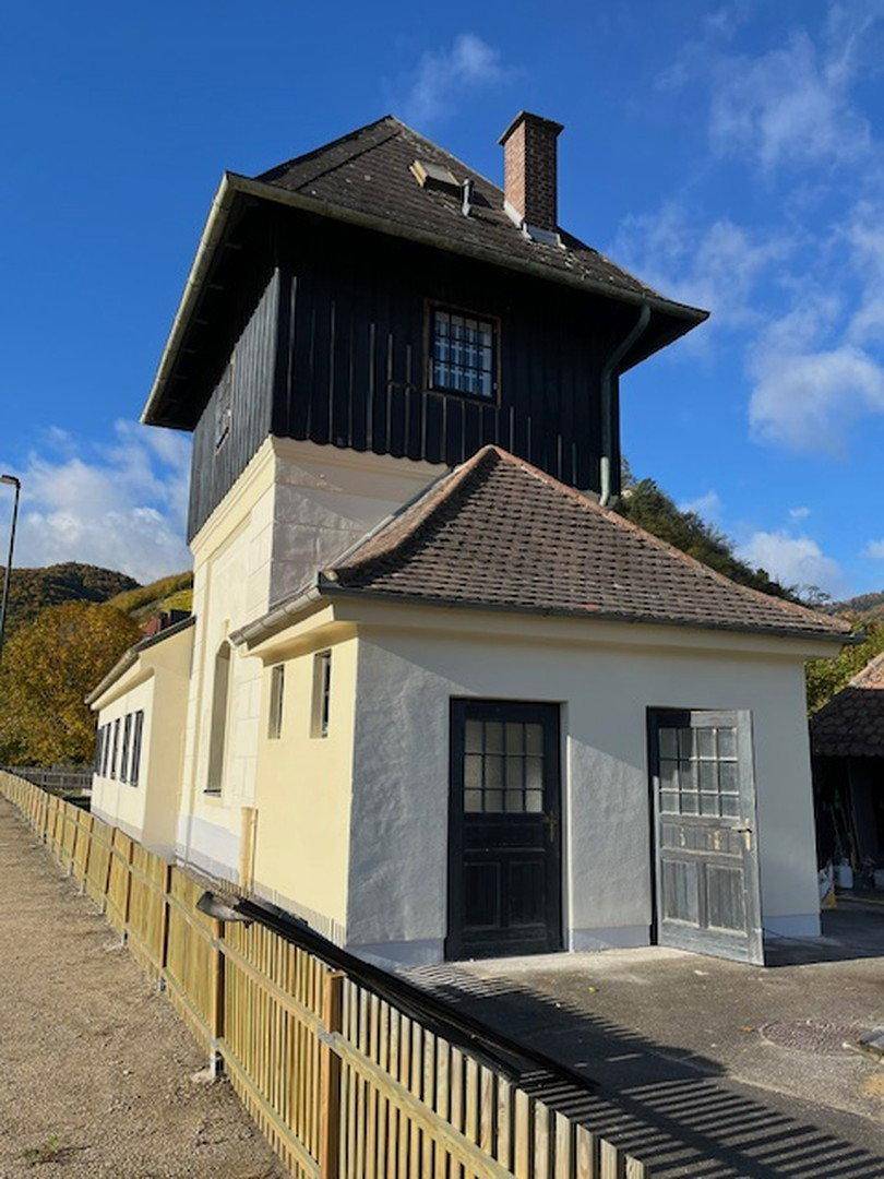 Historisches Gebäude mit markantem Turm und zwei Eingangstüren, umgeben von einer Holzumzäunung.