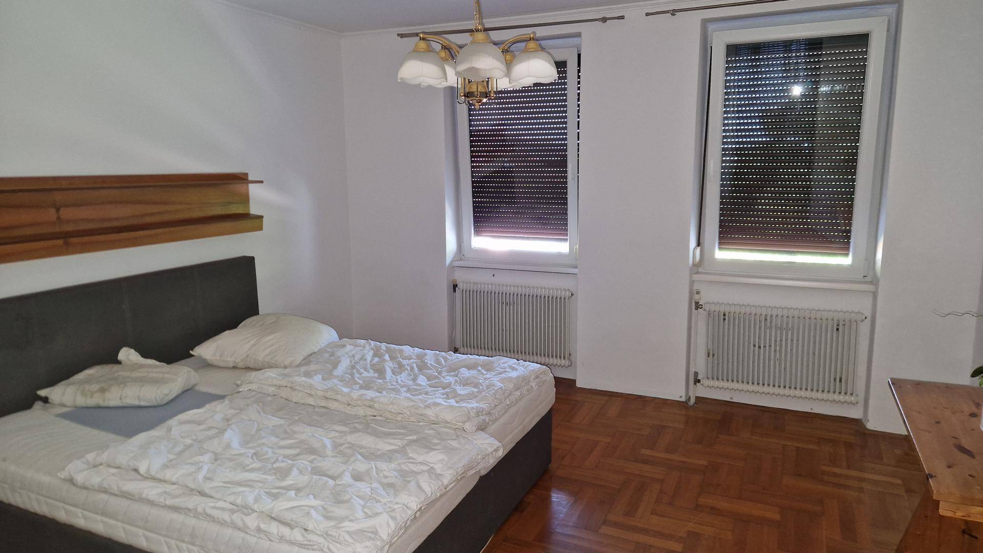 Schlafzimmer mit Doppelbett, Parkettboden und zwei Fenstern mit Rollläden.
