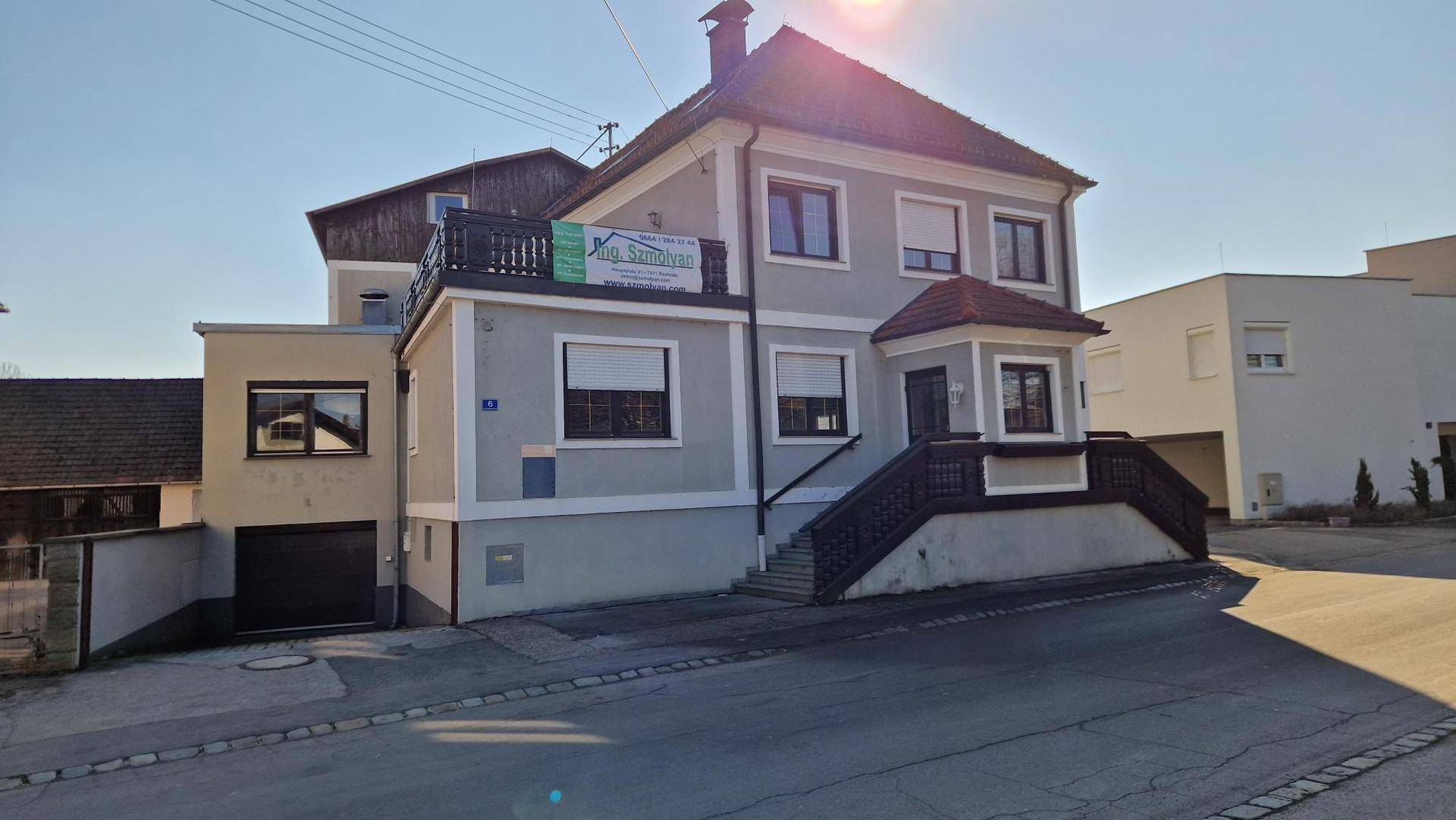 Graues Gebäude mit braunem Dach und Balkon, daneben eine Garage und ein gepflasterter Bereich.