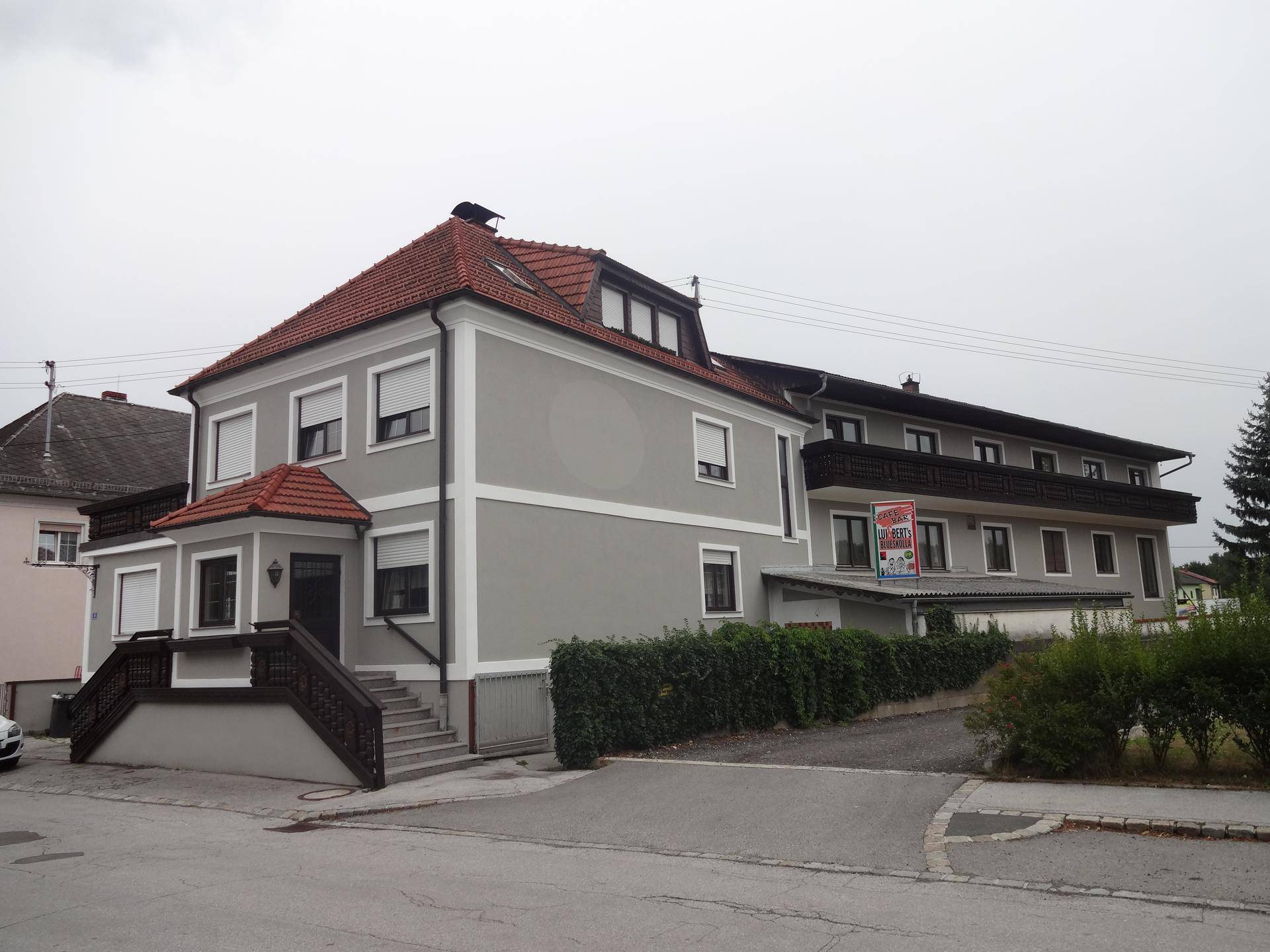 Graues Gebäude mit rotem Ziegeldach und Balkonen, umgeben von Bäumen und Sträuchern an einem bewölkten Tag.