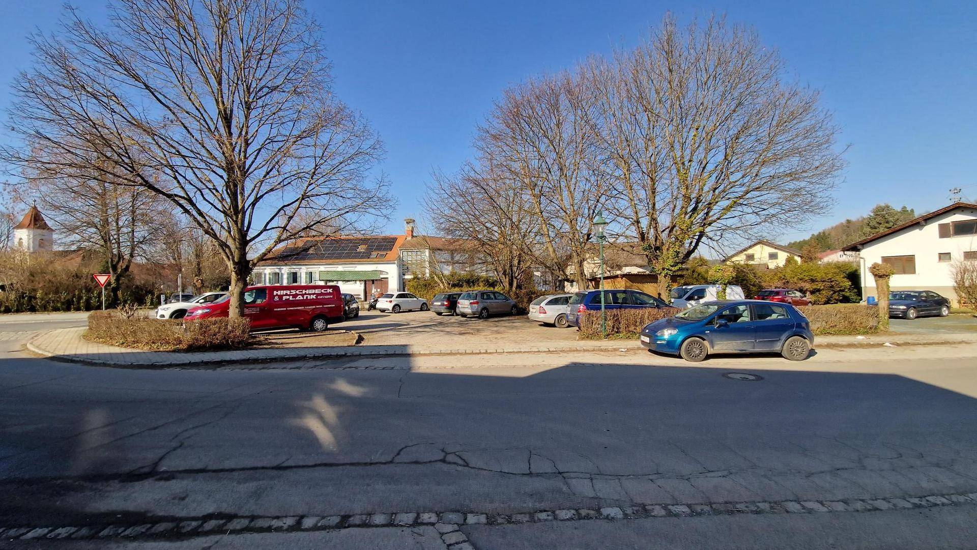 Weitläufiger Parkplatz mit mehreren Autos und großen Bäumen, im Hintergrund sind weitere Gebäude zu sehen.