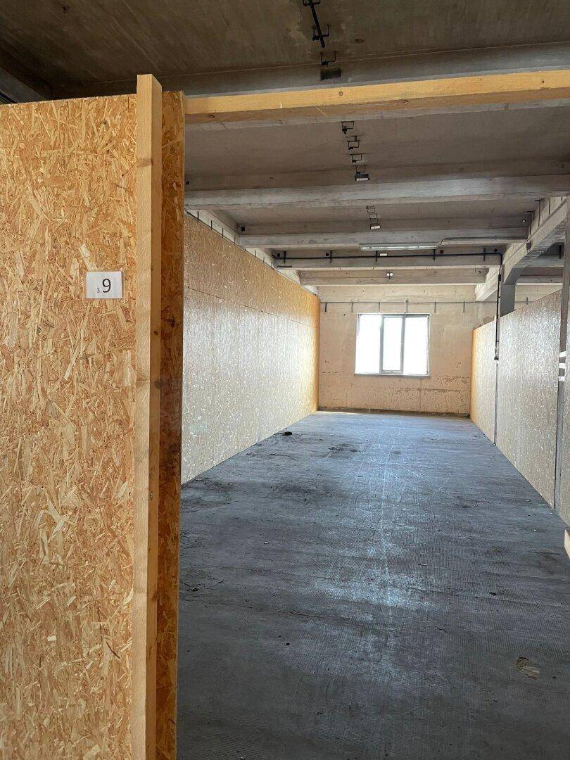 Langer, schmaler Lagerraum mit OSB-Wänden, Betonboden und einem kleinen Fenster am Ende.