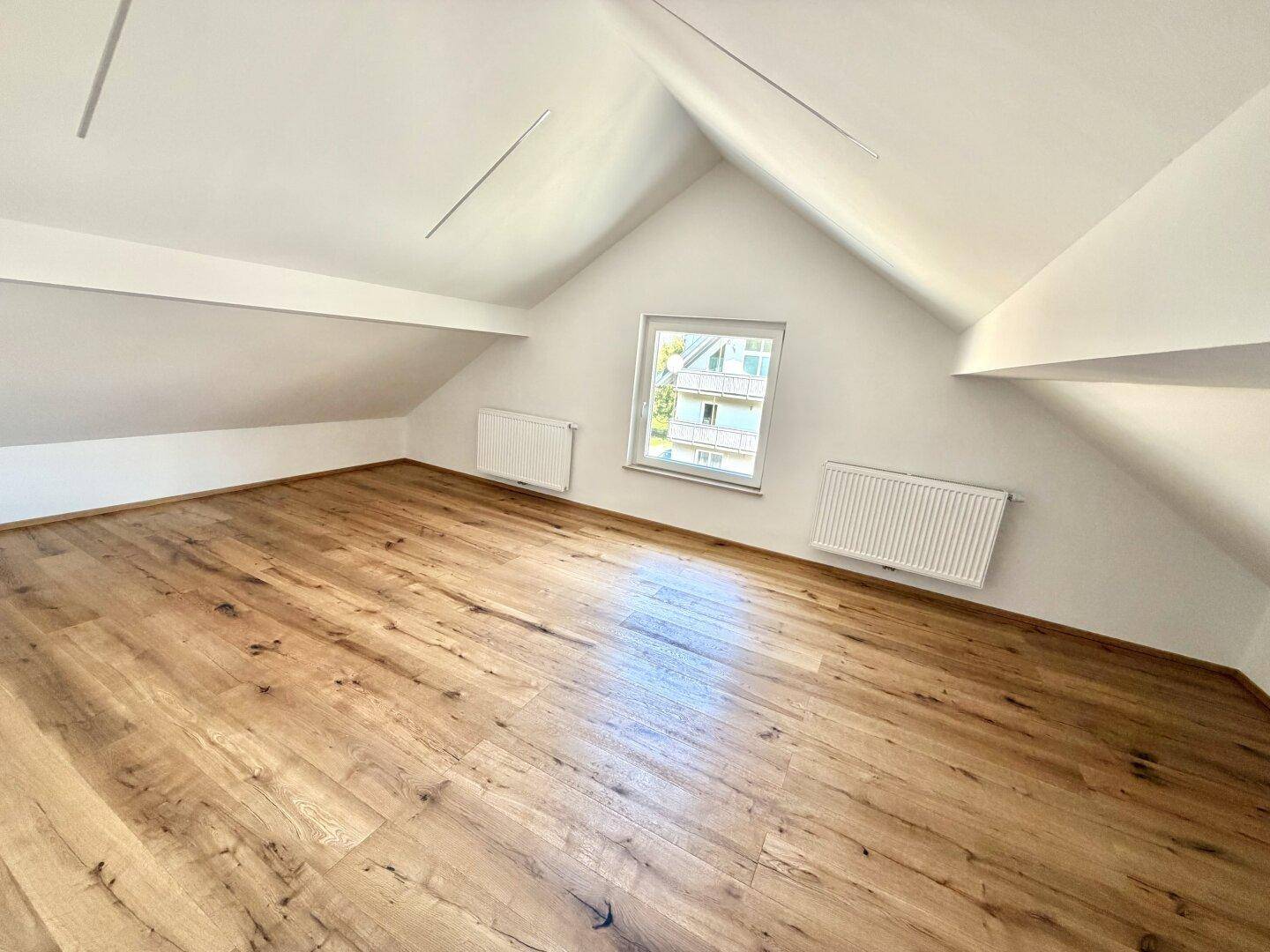 Geräumiges Dachzimmer mit Holzboden und schrägen Wänden, ideal als Schlaf- oder Arbeitsbereich.