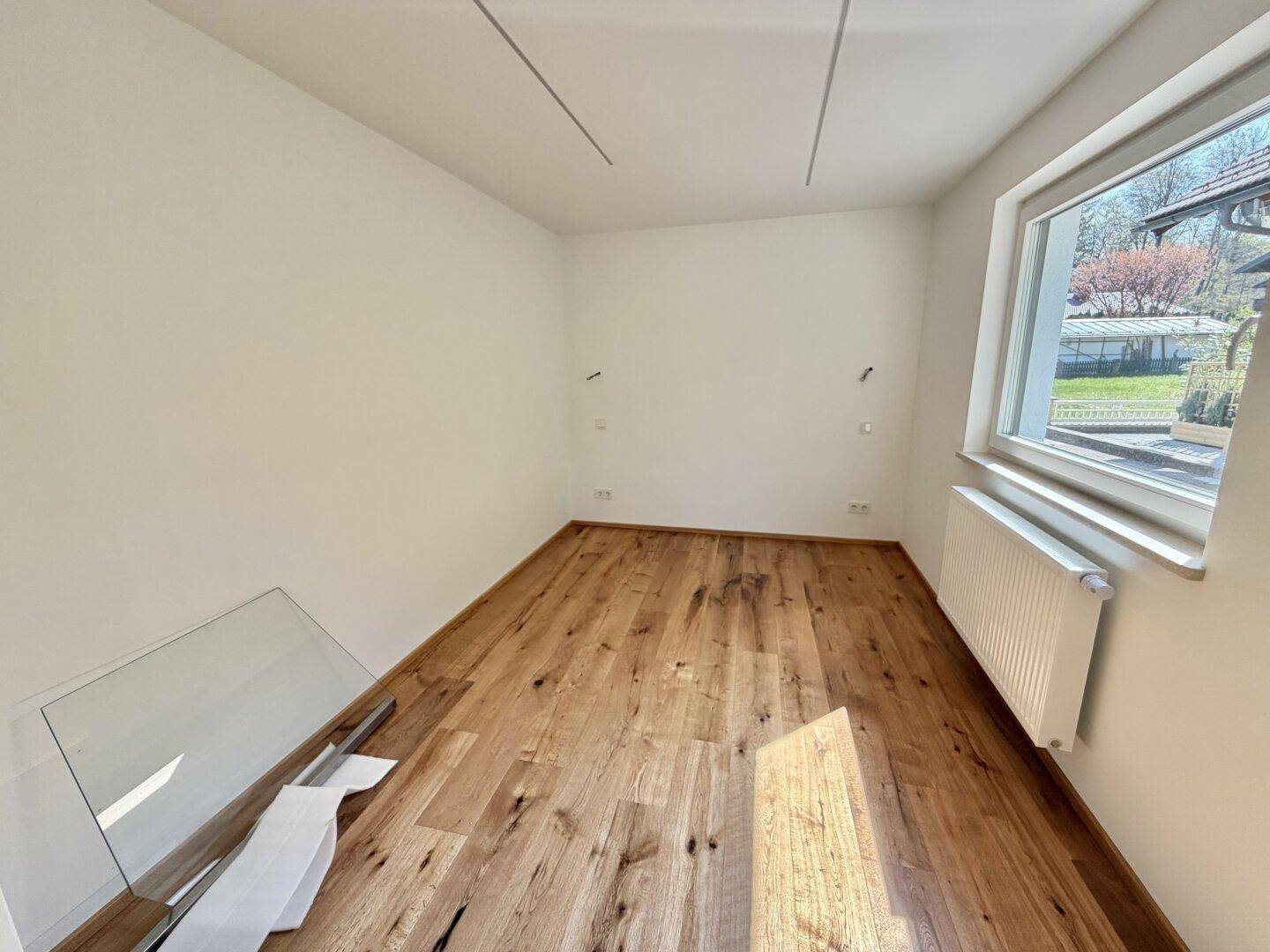 Freundliches Zimmer mit Holzboden und großem Fenster, das viel Tageslicht hereinlässt.