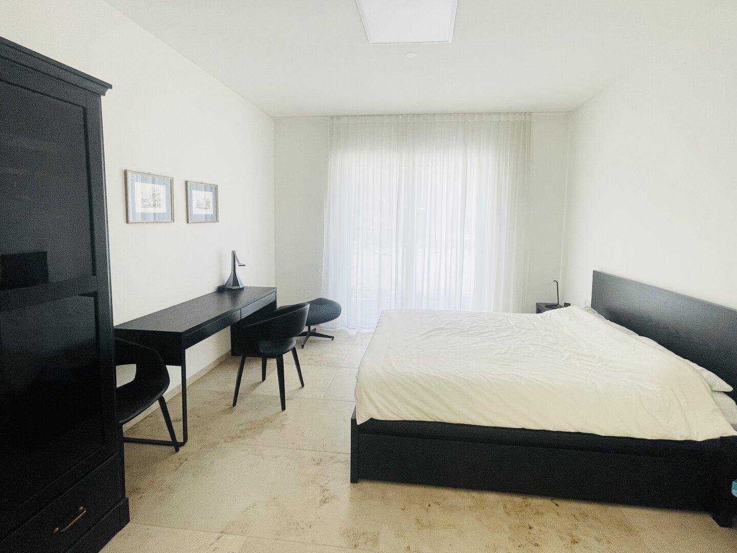 Modernes Schlafzimmer mit dunklem Bett, Schreibtisch und großem Fenster für viel Helligkeit.