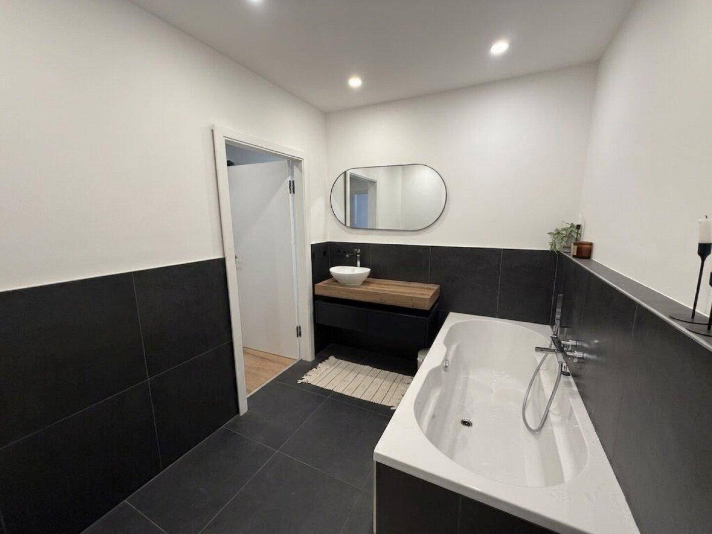 Helles Badezimmer mit Badewanne, modernem Holzwaschtisch und dunklen Wand- und Bodenfliesen.