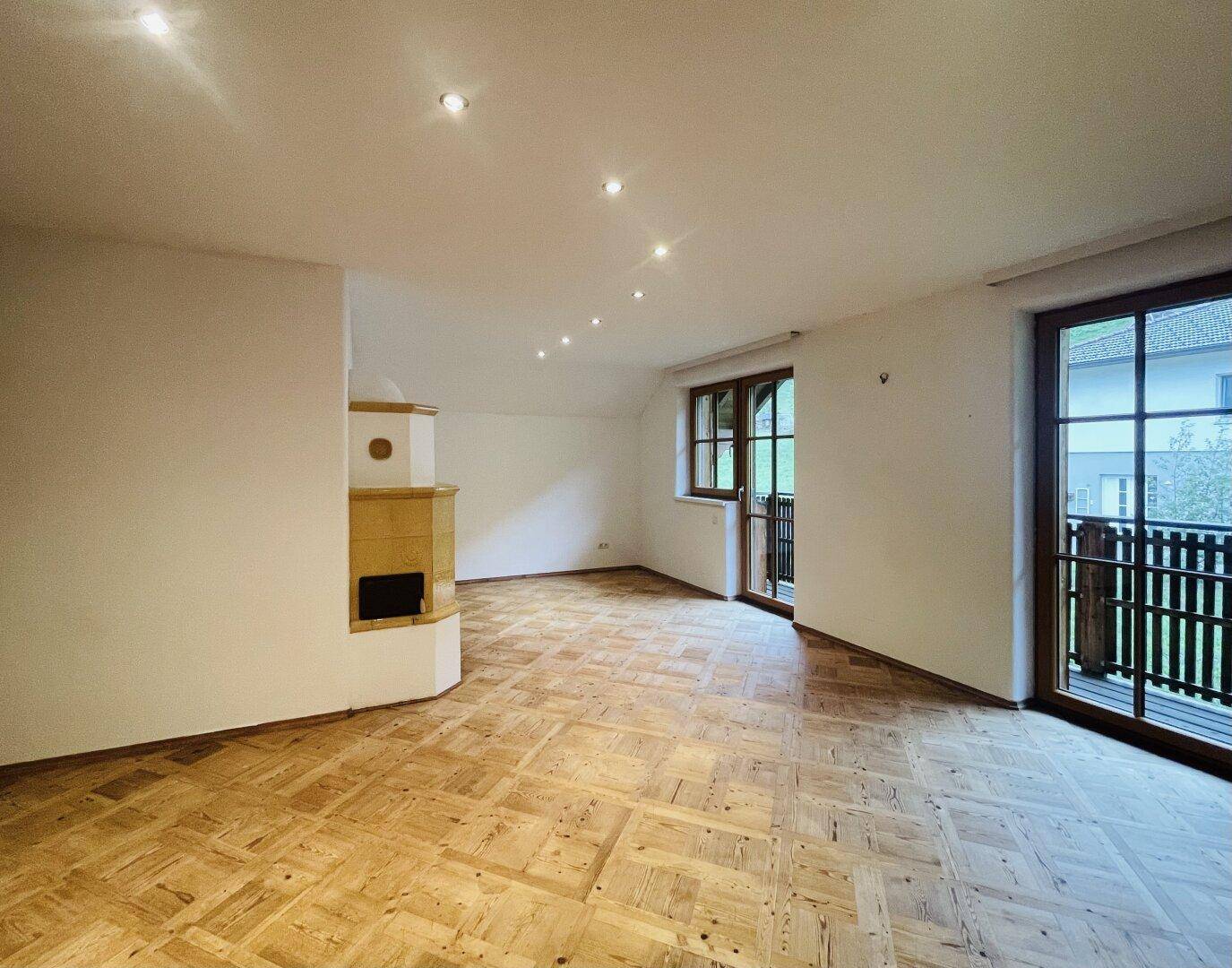 Geräumiges Wohnzimmer mit traditionellem Kachelofen, Parkettboden und Zugang zum Balkon.
