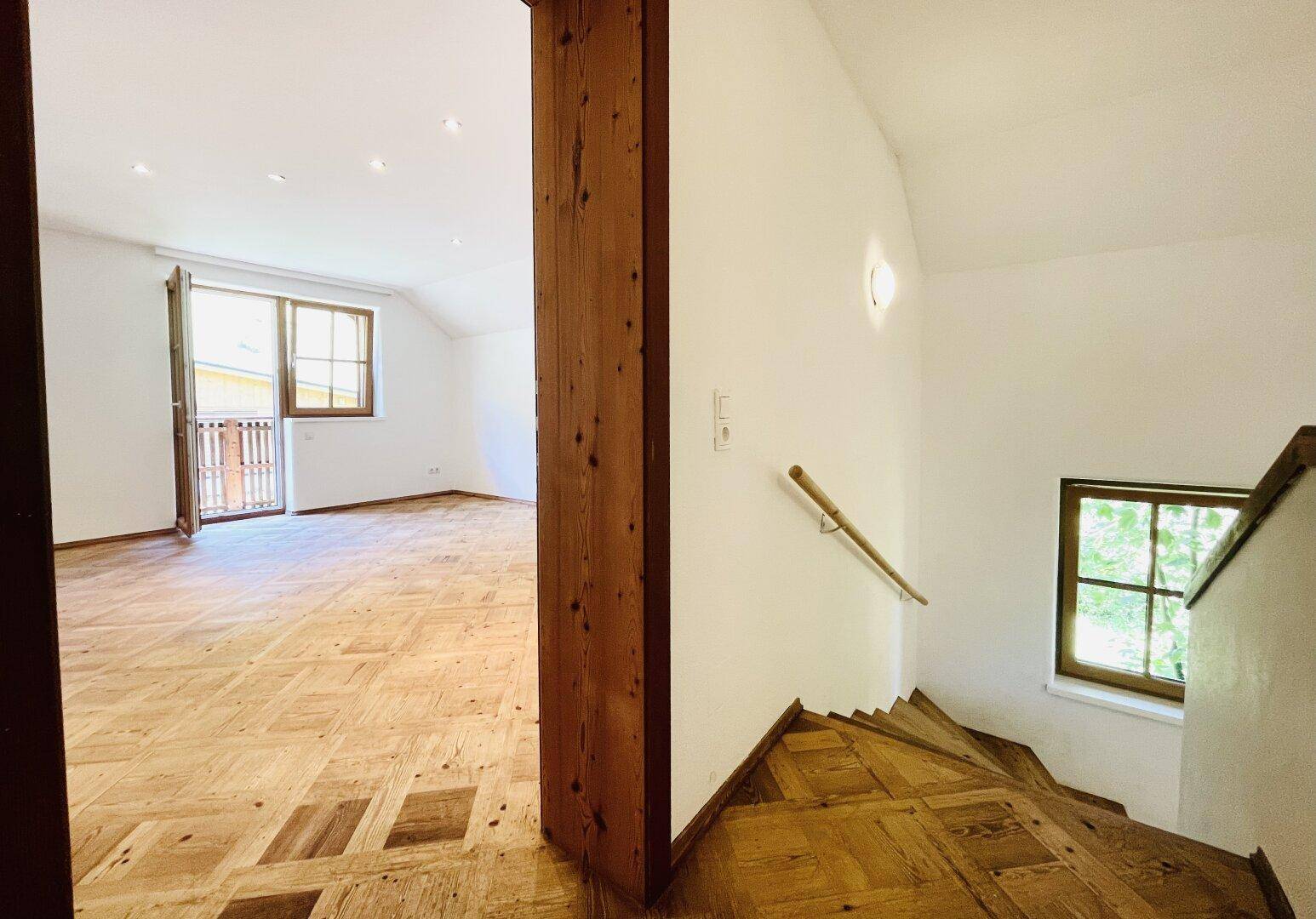 Heller Innenbereich mit charakteristischem Holzparkettboden, Holztreppe und Zugang zu einem Balkon.