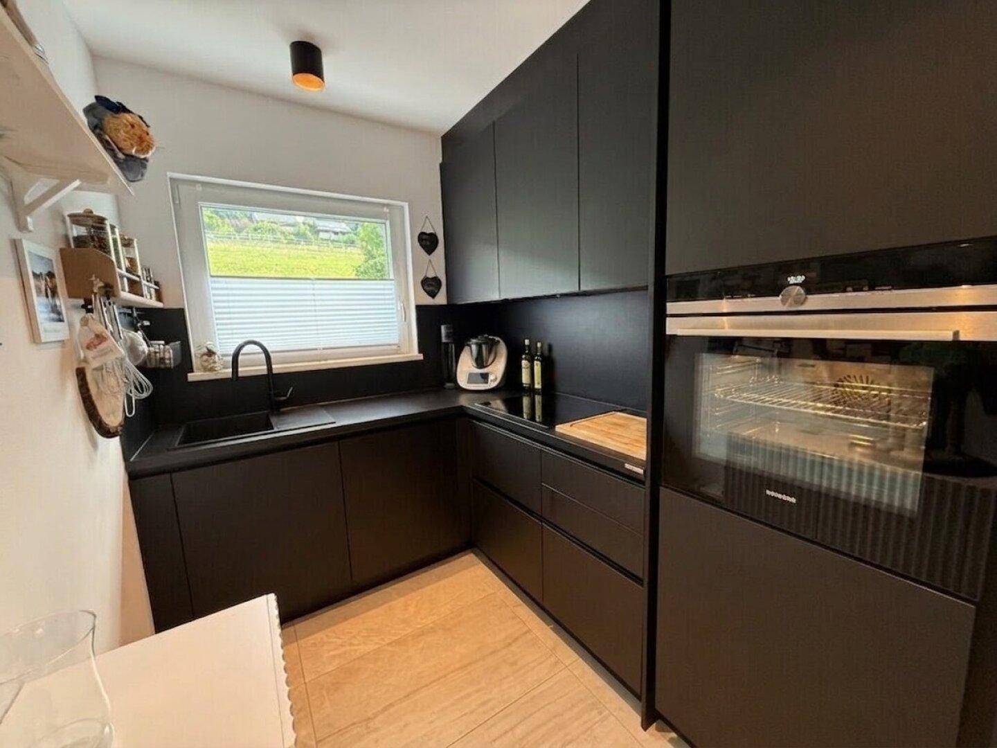 Moderne schwarze Küche mit integrierten Geräten, Induktionskochfeld und Fensterblick ins Grüne.
