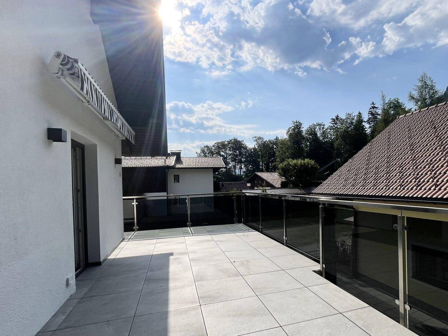 Sonnige Terrasse mit modernen Fliesen, Glasgeländer und weitem Blick auf die grüne Umgebung.