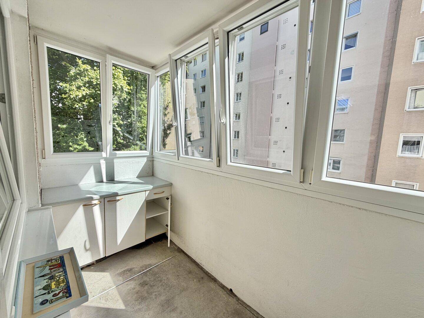 Verglaster Balkon mit Blick auf umliegende Gebäude und Bäume, ausgestattet mit einem kleinen Schrank.