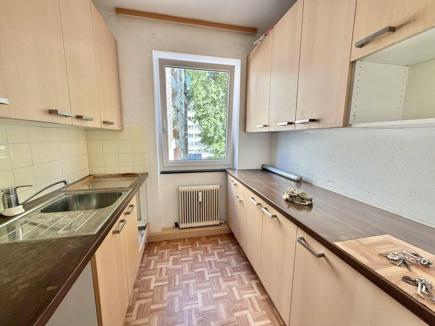 Schmale Küche mit Holzschränken, Spüle und Fenster mit Blick ins Grüne.