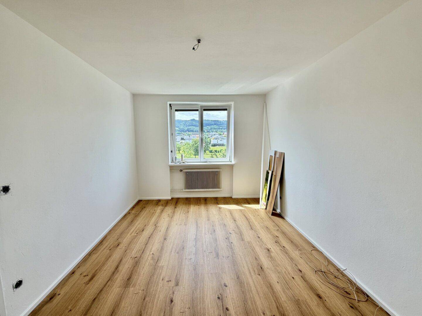 Leeres Schlafzimmer mit Laminatboden und einem großen Fenster, das viel Tageslicht hereinlässt.