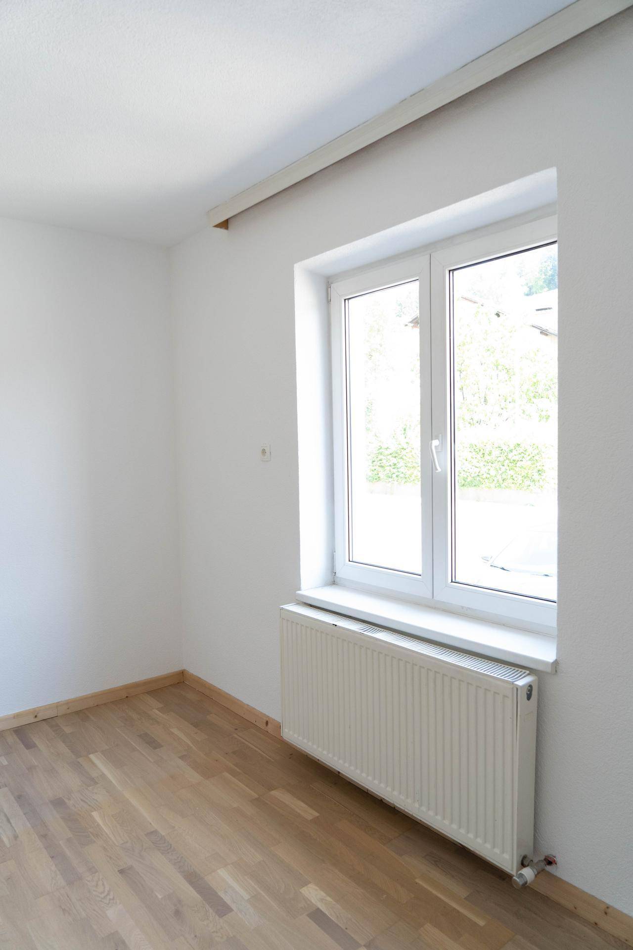 Helles, leeres Zimmer mit einem Fenster, Parkettboden und Heizkörper, bereit zur Einrichtung.