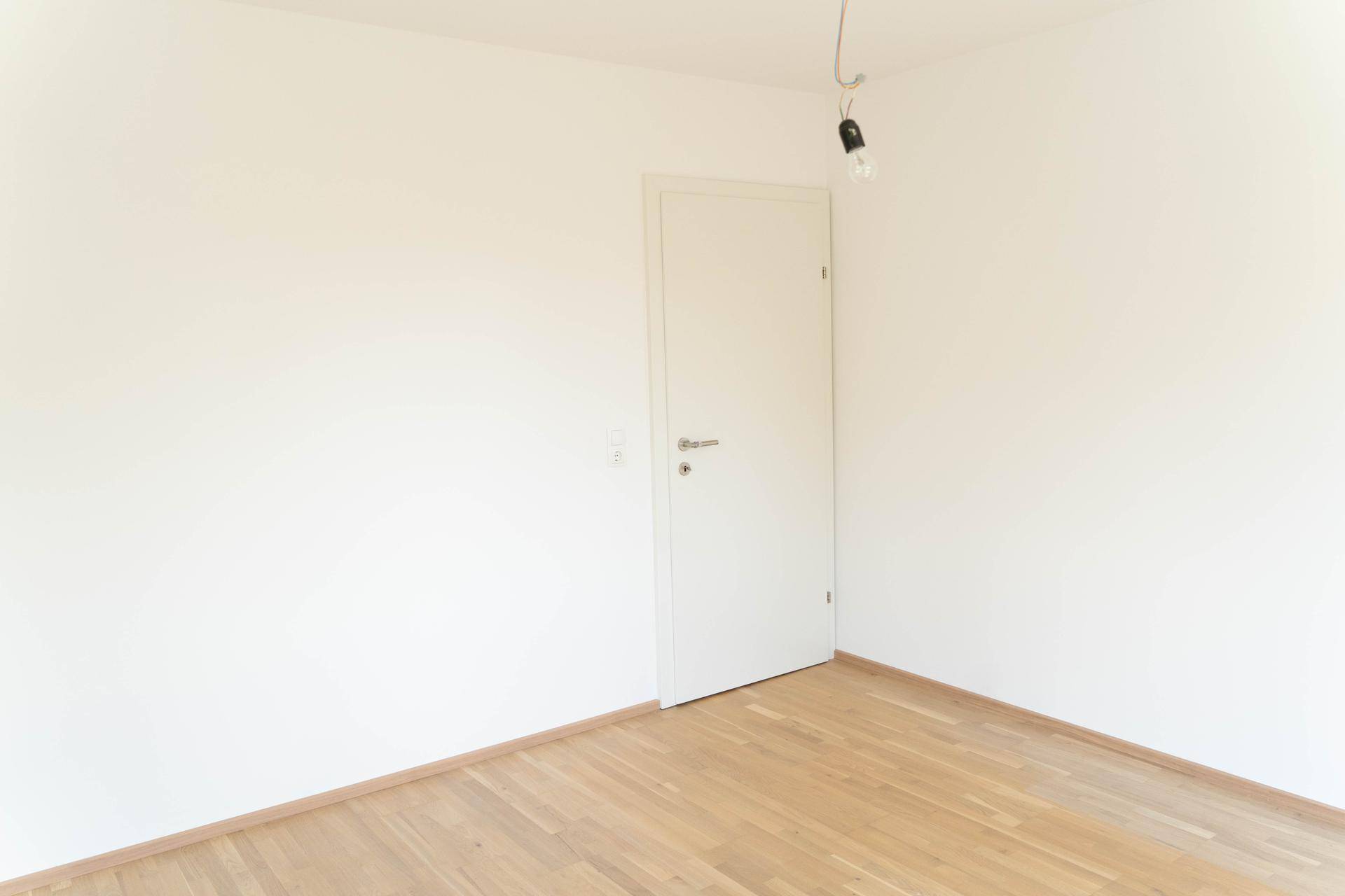 Leere Zimmerecke mit weißer Tür und hellem Parkettboden, schlicht und funktional.