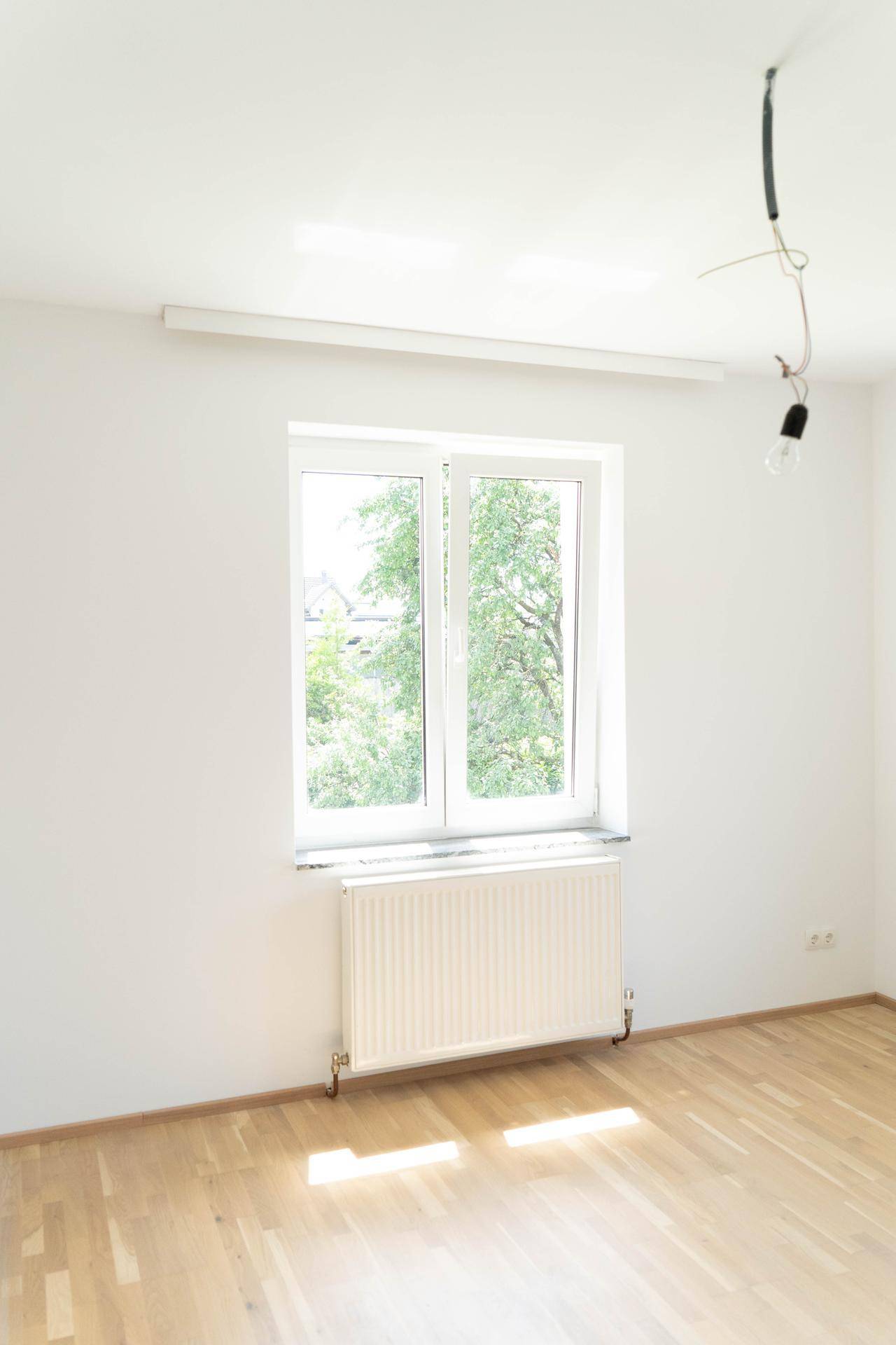 Lichtdurchflutetes Zimmer mit Parkettboden und großem Fenster, das eine grüne Aussicht bietet.