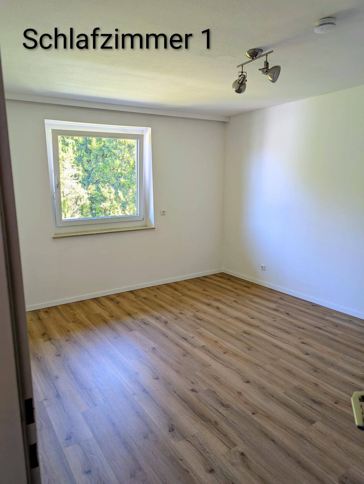 Helles Schlafzimmer mit Laminatboden und einem Fenster, das einen angenehmen Blick ins Grüne bietet.