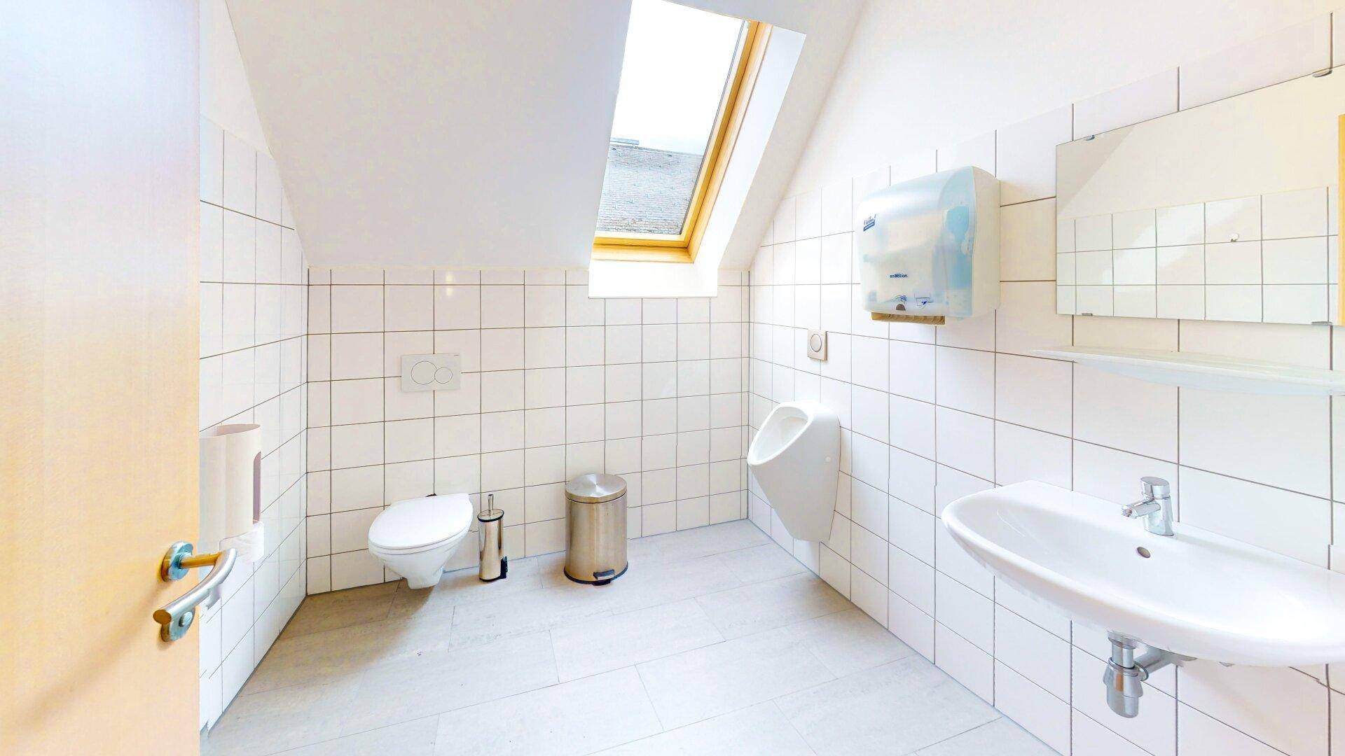 WC-Raum mit weißen Fliesen, Toilette, Urinal und Dachfenster für Belüftung.