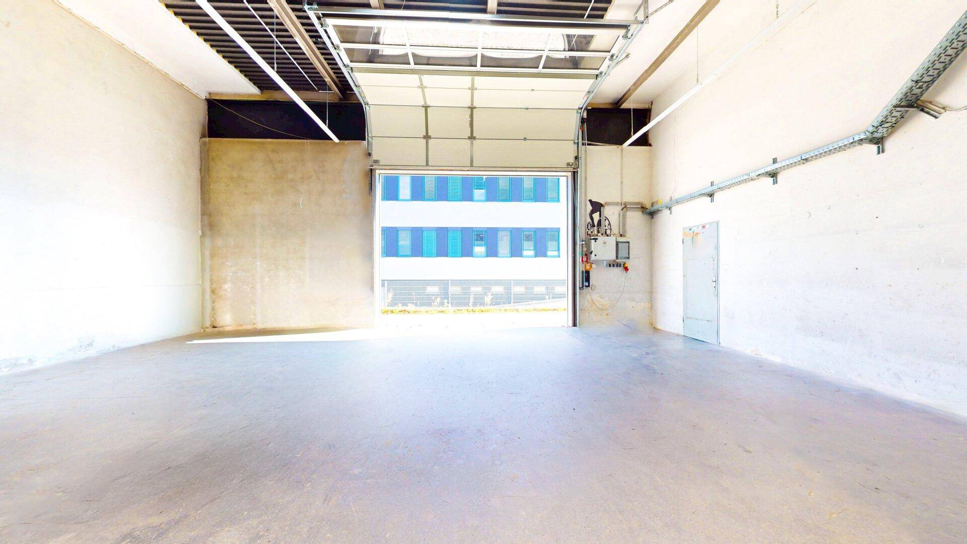 Leere Lagerhalle mit offenem Rolltor, das den Blick auf ein gegenüberliegendes Gebäude und den Außenbereich freigibt.