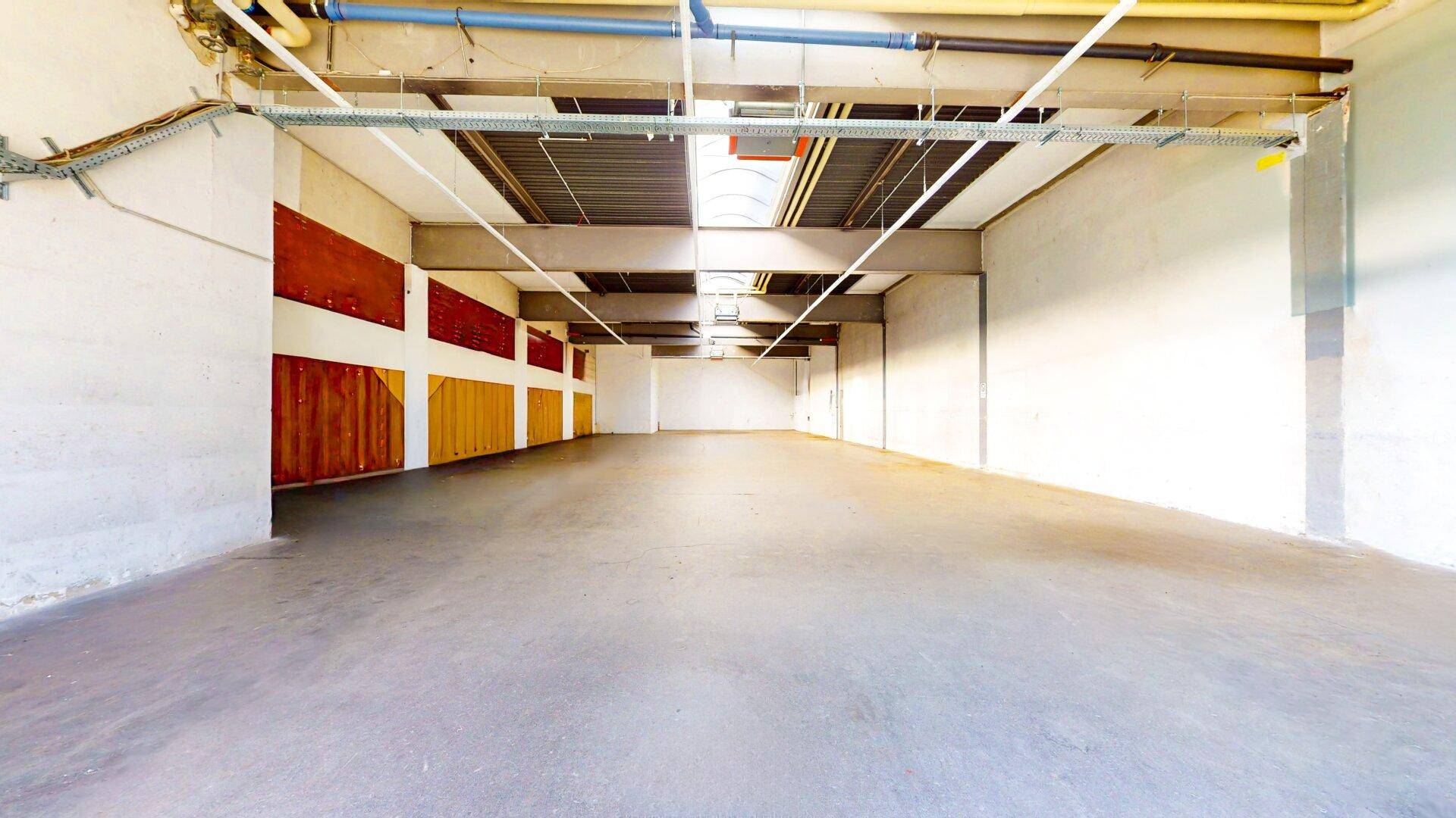 Leere Industriehalle mit Betonboden, hohen Decken, sichtbaren Leitungen und Oberlichtern für natürliches Licht.