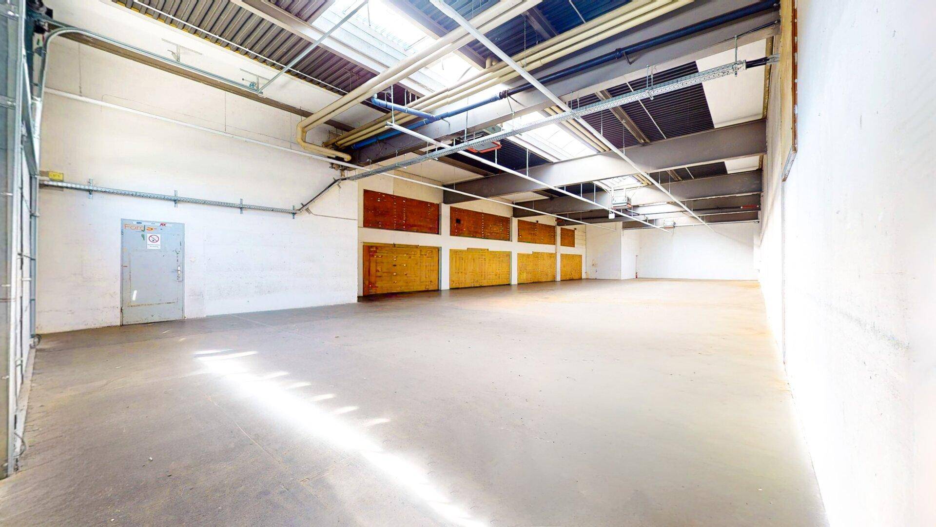 Große, leere Lagerhalle mit Betonboden, hohen Decken, Oberlichtern und einem großen Rolltor am Eingang.