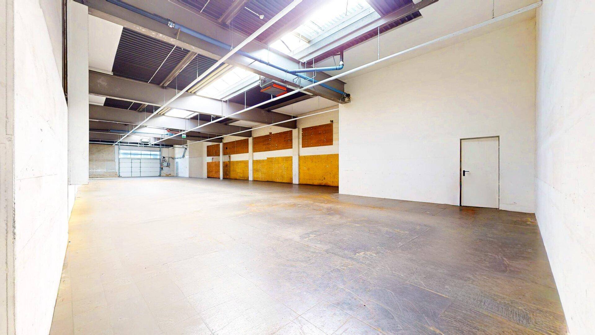 Geräumige, leere Lagerhalle mit Betonboden, hohen Decken, Oberlichtern und einem Rolltor am Ende des Raumes.