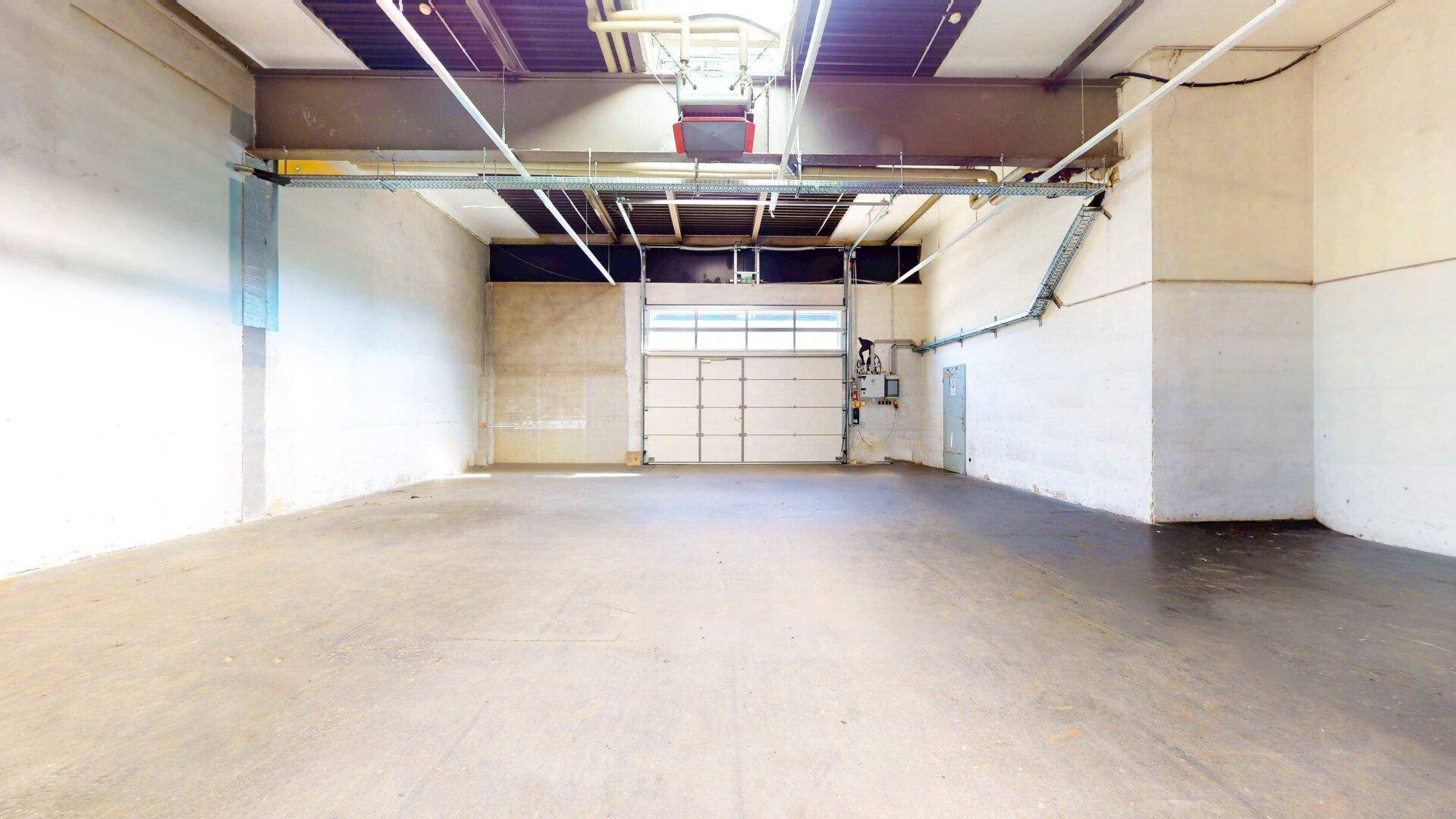 Geräumige, leere Lagerhalle mit Betonboden, hohen Decken, Oberlichtern und einem geschlossenen Rolltor am Ende.