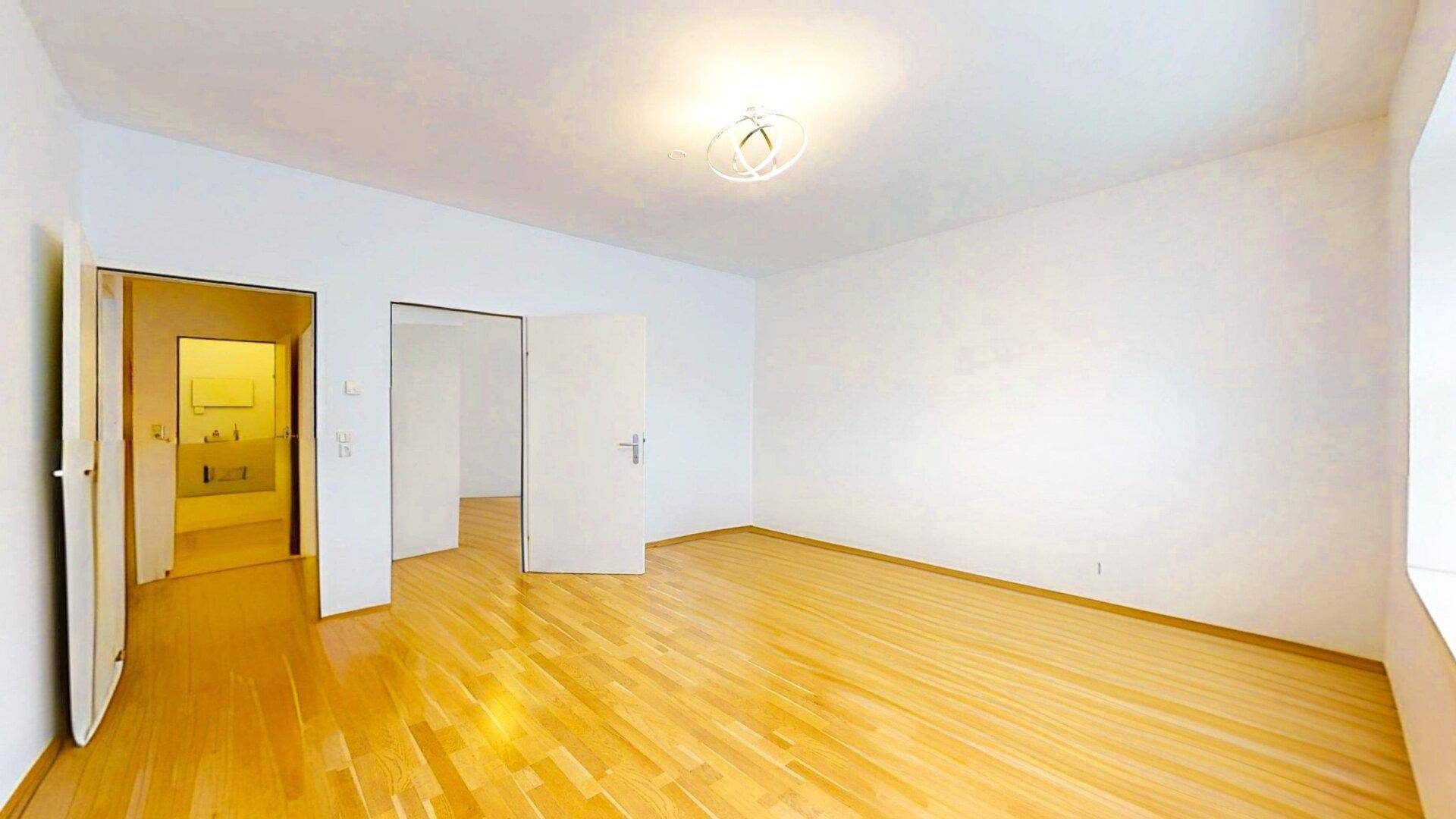 Geräumiges, leeres Zimmer mit hellem Holzparkett und weißen Wänden, mit Blick auf das Badezimmer.
