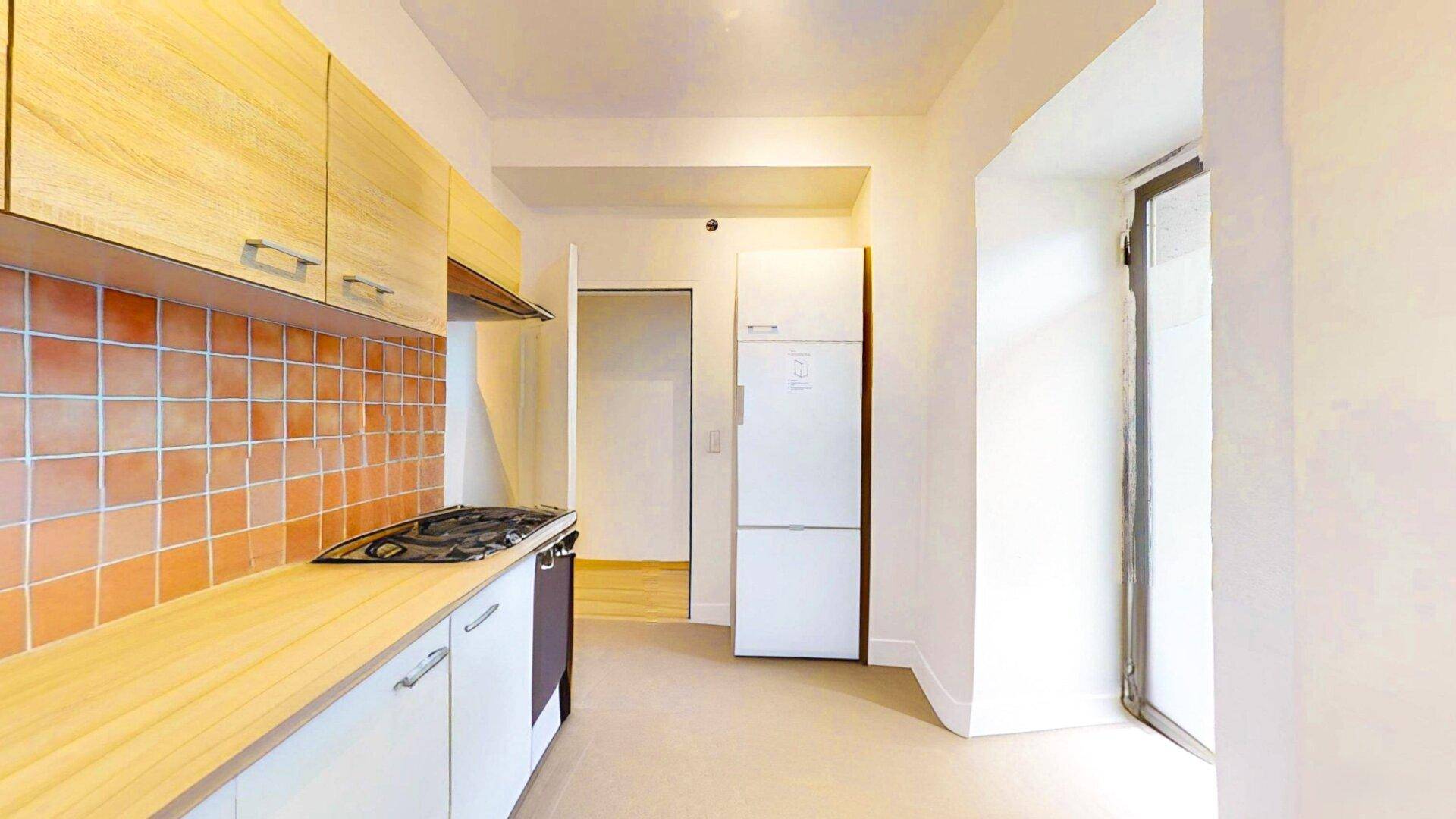 Kleine Küche mit Holzschränken, Kühlschrank und Fenster, bietet grundlegende Kochmöglichkeiten.