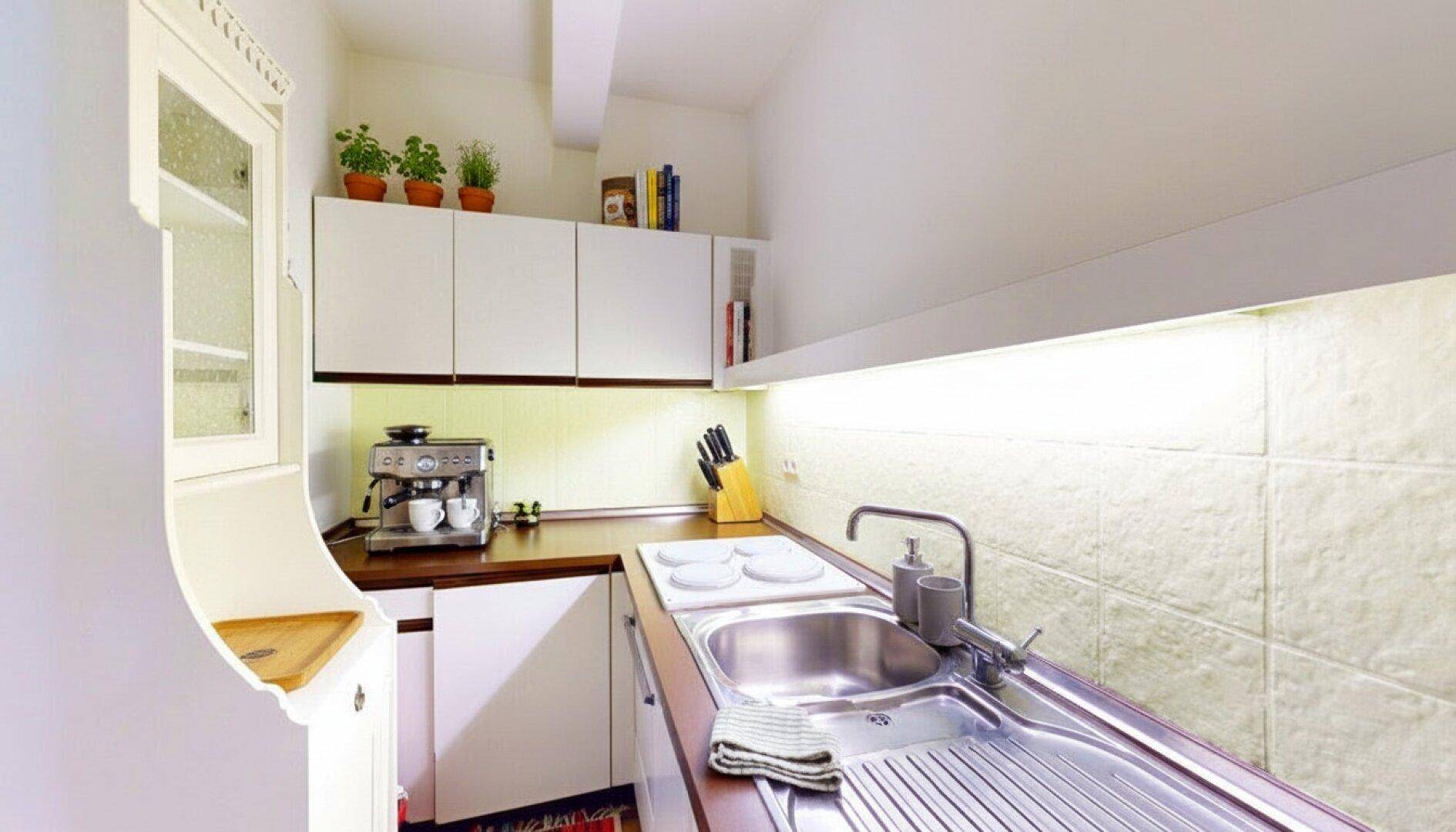 Kleine, funktionale Küche mit weißen Schränken, Edelstahlspüle und Kaffeemaschine.