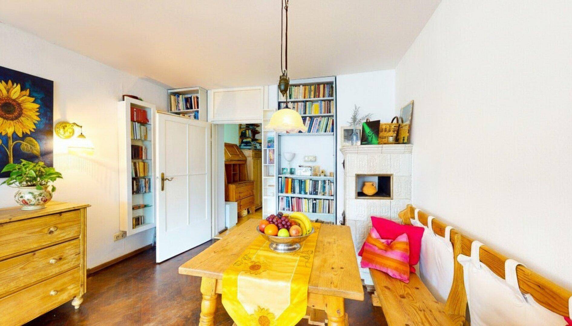 Gemütlicher Essbereich mit Holztisch, Sitzbank und Bücherregalen, ergänzt durch einen Kachelofen.