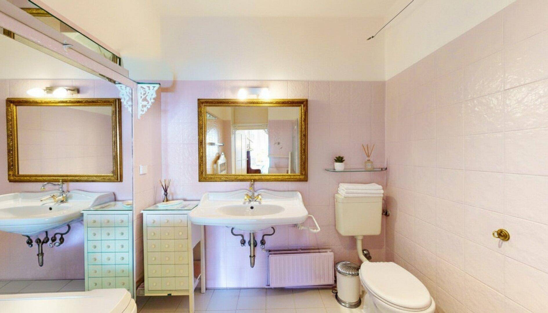 Badezimmer im 80er-Jahre-Stil mit rosa Fliesen, zwei Waschbecken und Toilette.