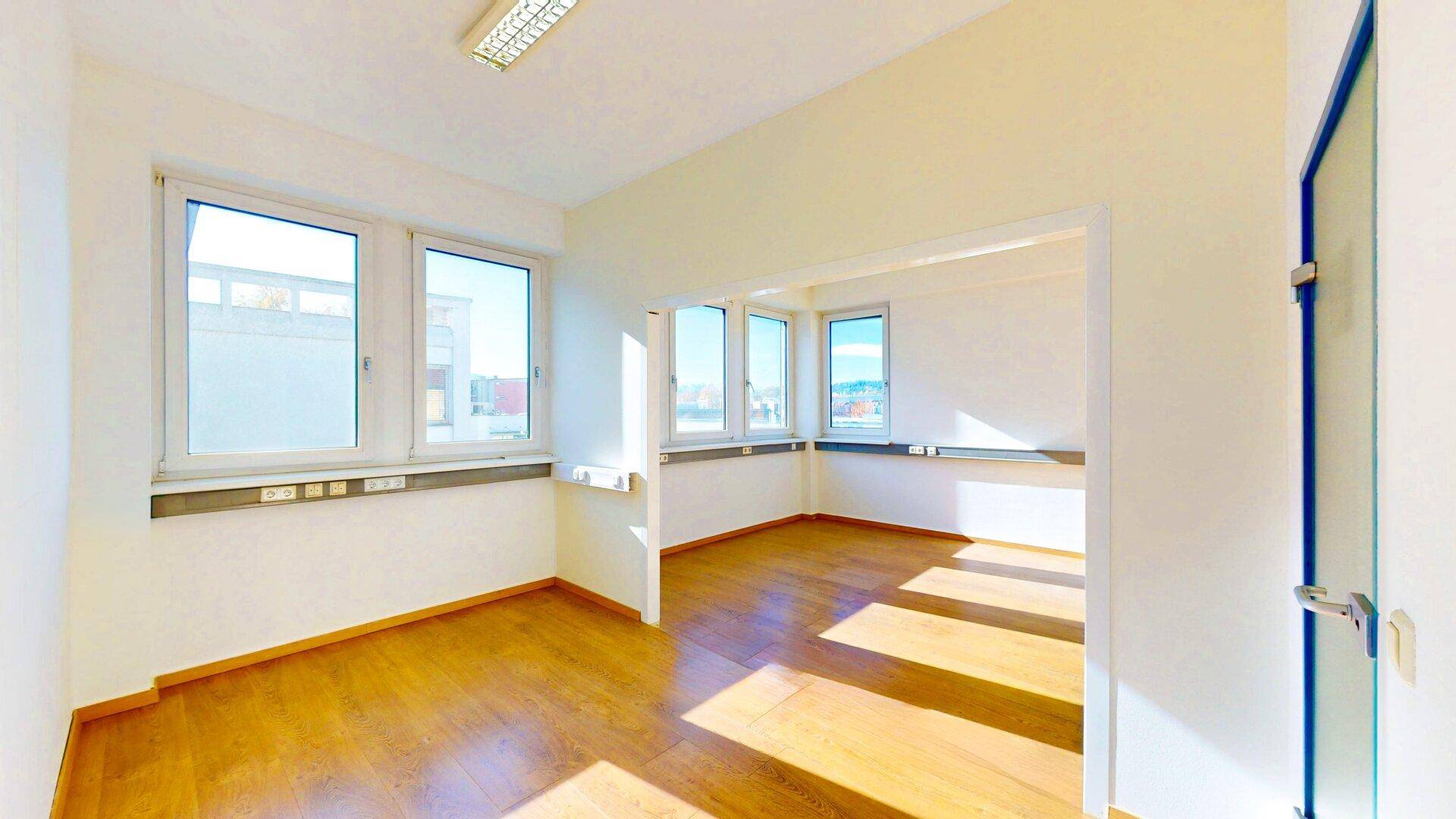 Zwei miteinander verbundene, leere Büroräume mit großen Fenstern, die viel Tageslicht hereinlassen, und Holzfußboden.