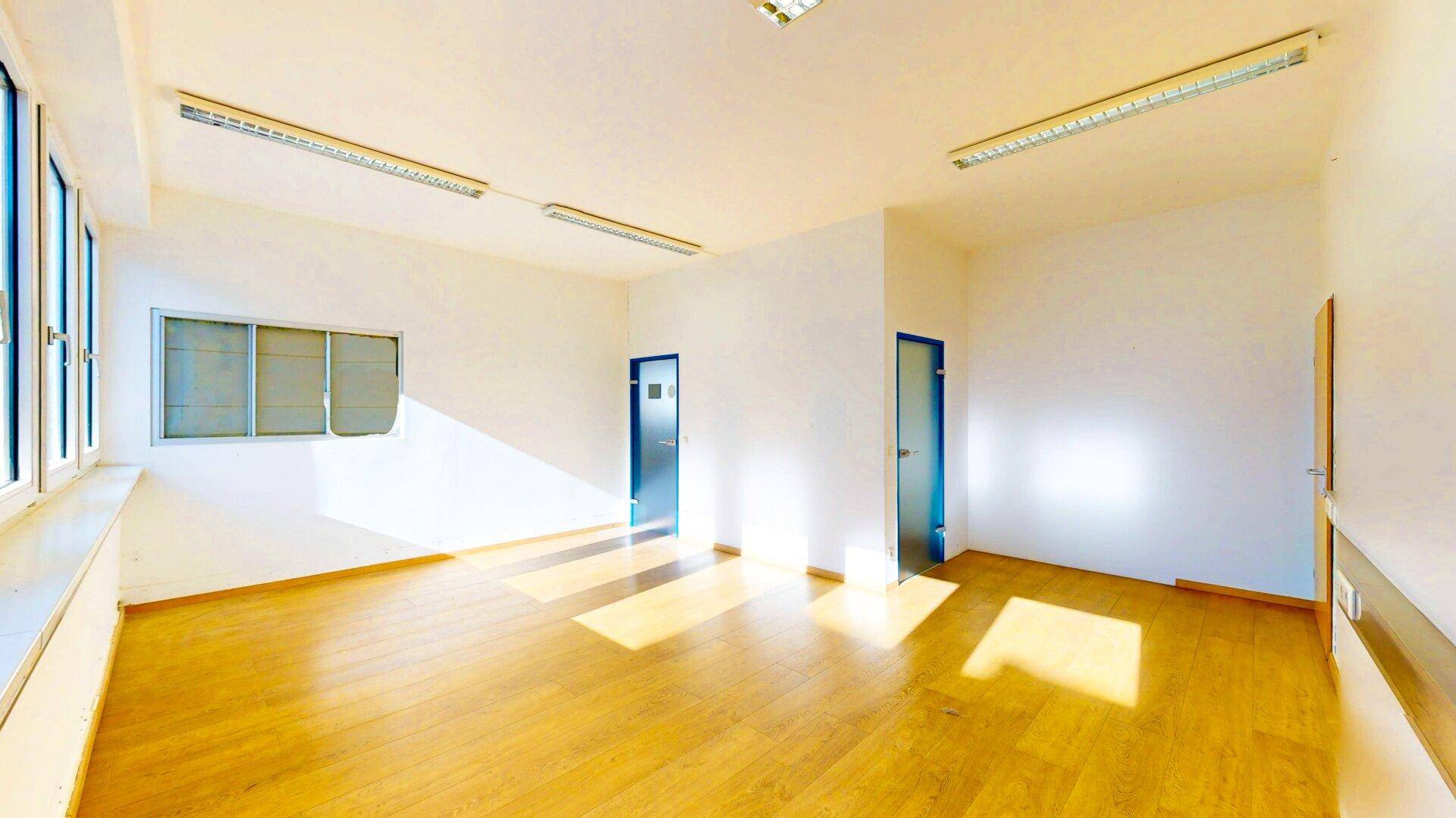 Heller, leerer Büroraum mit großen Fenstern und zwei Türen, die zu weiteren Räumen führen, mit viel Sonnenlicht.