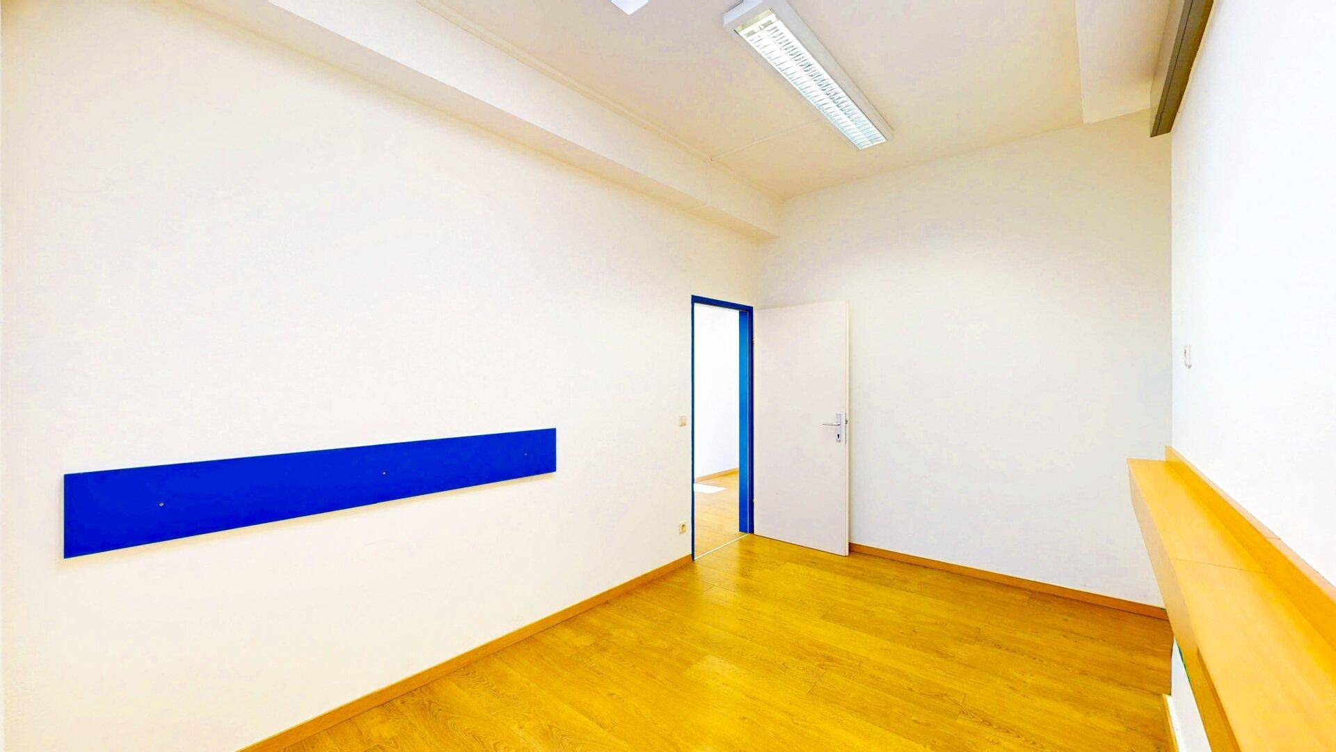 Leerer Büroraum mit hellem Holzfußboden und einer Tür, die zu einem weiteren Raum führt, mit blauer Akzentleiste.