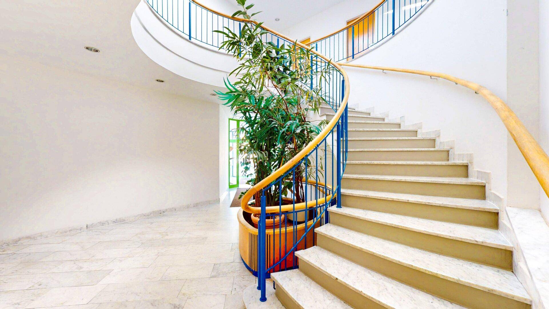 Elegantes Treppenhaus mit geschwungener Marmortreppe, blauem Geländer und Pflanzen, hell und offen.