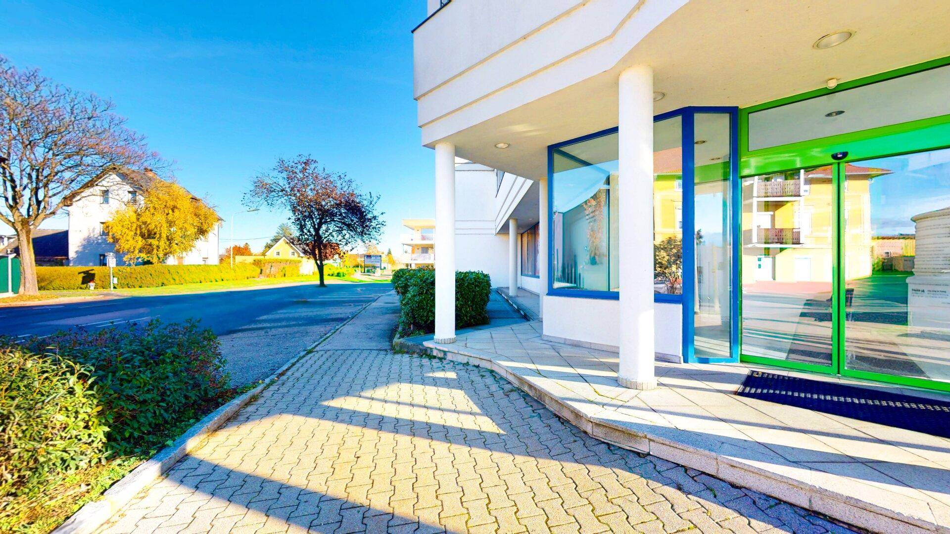 Gebäudeeingang mit Säulen und Glasfassade, daneben ein Bürgersteig und eine Straße mit Bäumen im Herbst.
