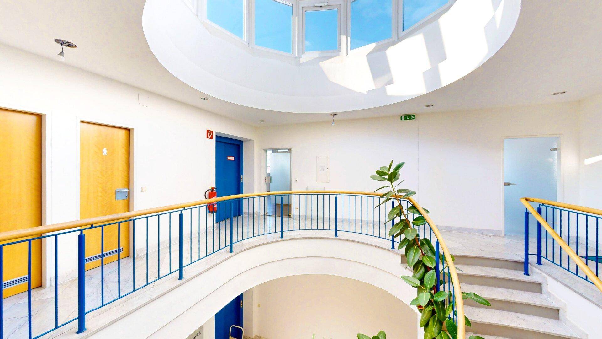 Helles Treppenhaus mit runder Oberlichtkuppel, blauen Geländern und mehreren Türen, die zu verschiedenen Räumen führen.
