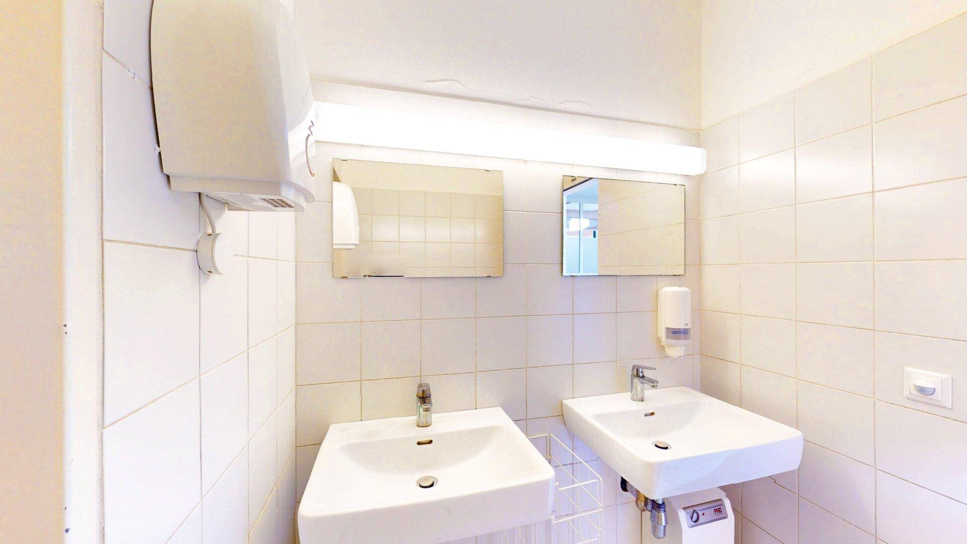 Heller Waschraum mit zwei Waschbecken, weißen Fliesen und einem Handtrockner, sauber und funktional gestaltet.