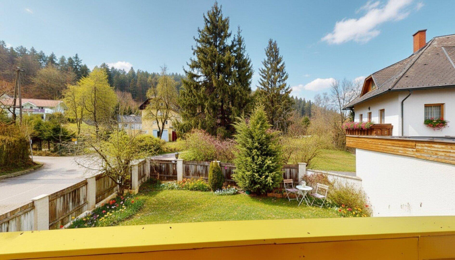 Blick vom Balkon auf den grünen Garten und die umliegende, bewaldete Landschaft.