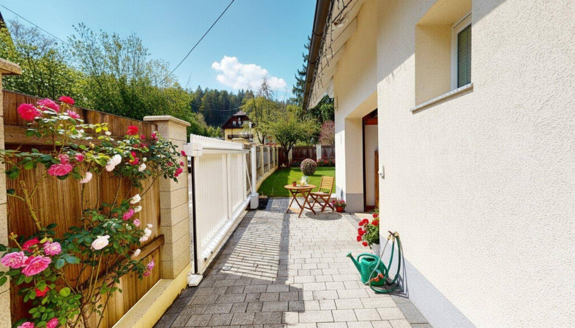 Gepflasterter Weg entlang des Hauses mit blühenden Rosen und einem kleinen Sitzbereich im Garten.