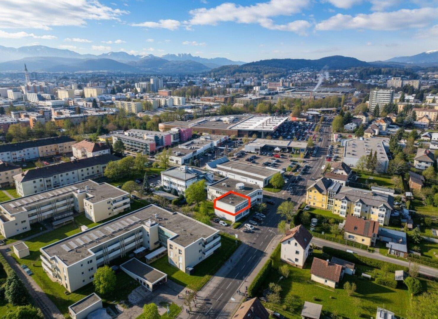 Weitläufige Luftaufnahme einer Stadt mit Bergen im Hintergrund und einem markierten Gebäude im Zentrum.